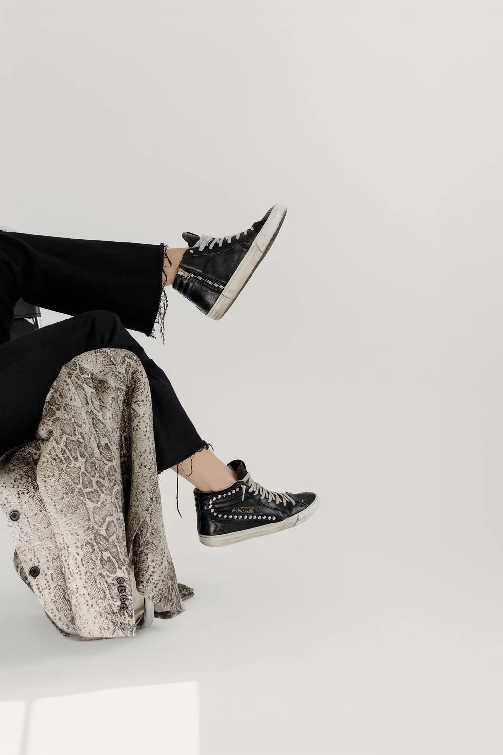 Close-up of a person's legs and feet wearing black high-top sneakers with silver embellishments and white soles, with a light-colored snakeskin patterned jacket draped over their legs, against a plain white background.
