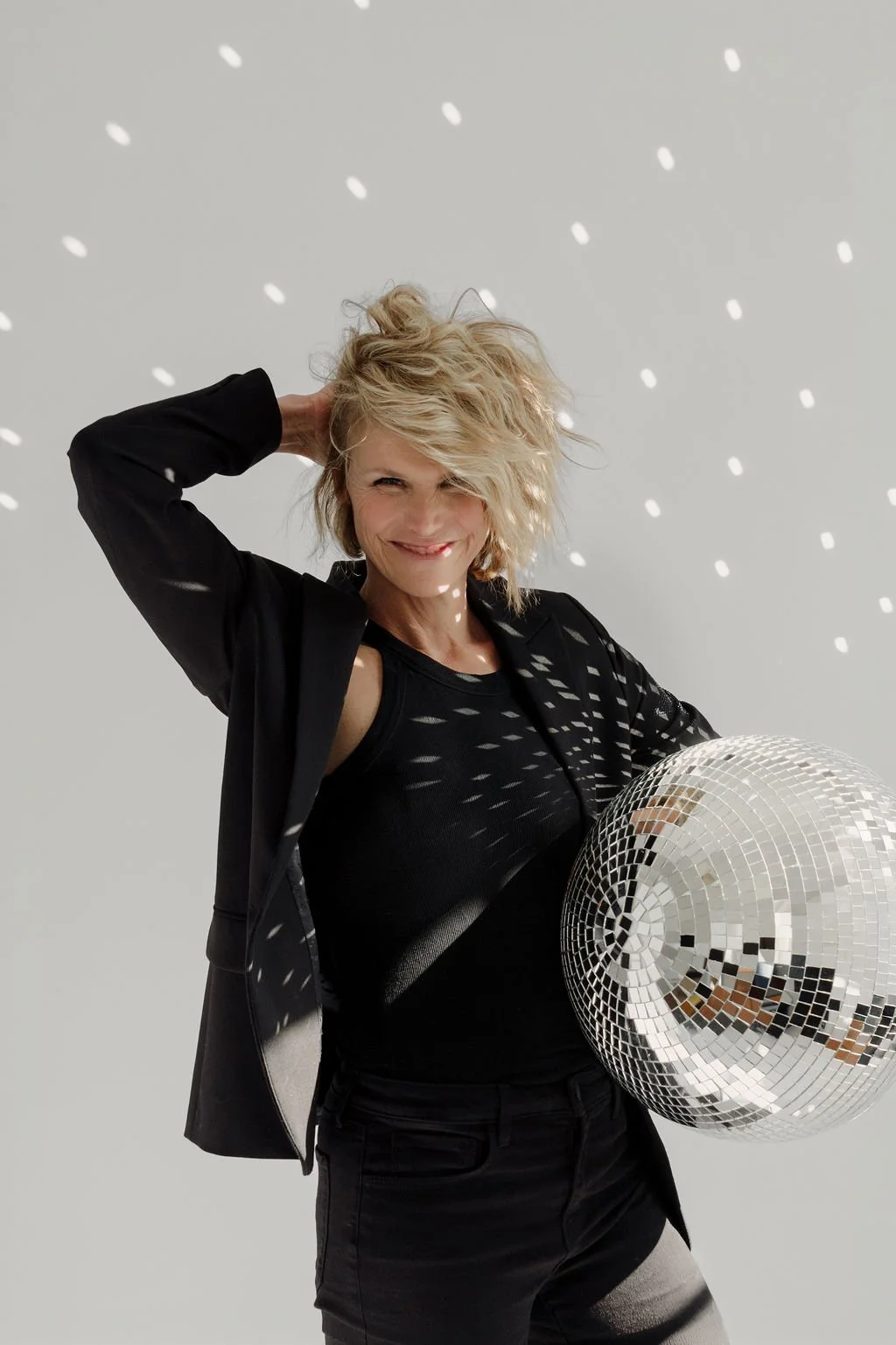 Woman with blonde curly hair holding a disco ball, with white dots of light reflecting off the ball and background.