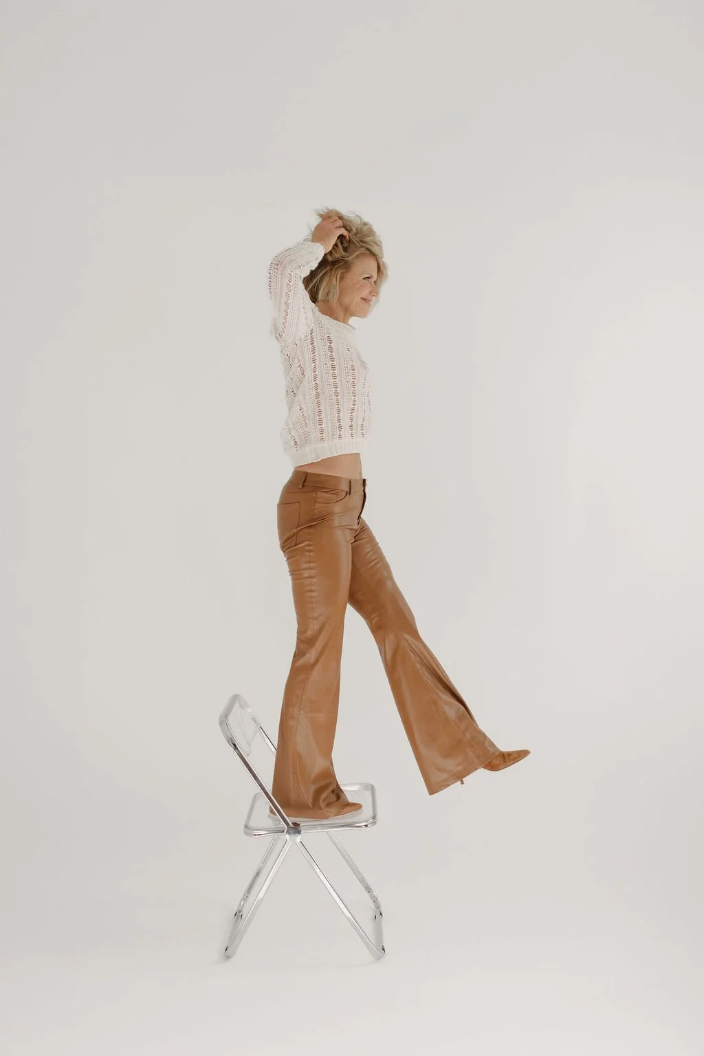 A woman standing on a clear plastic chair, wearing a white knitted sweater and brown leather pants, with one hand running through her blonde hair, smiling against a plain white background.