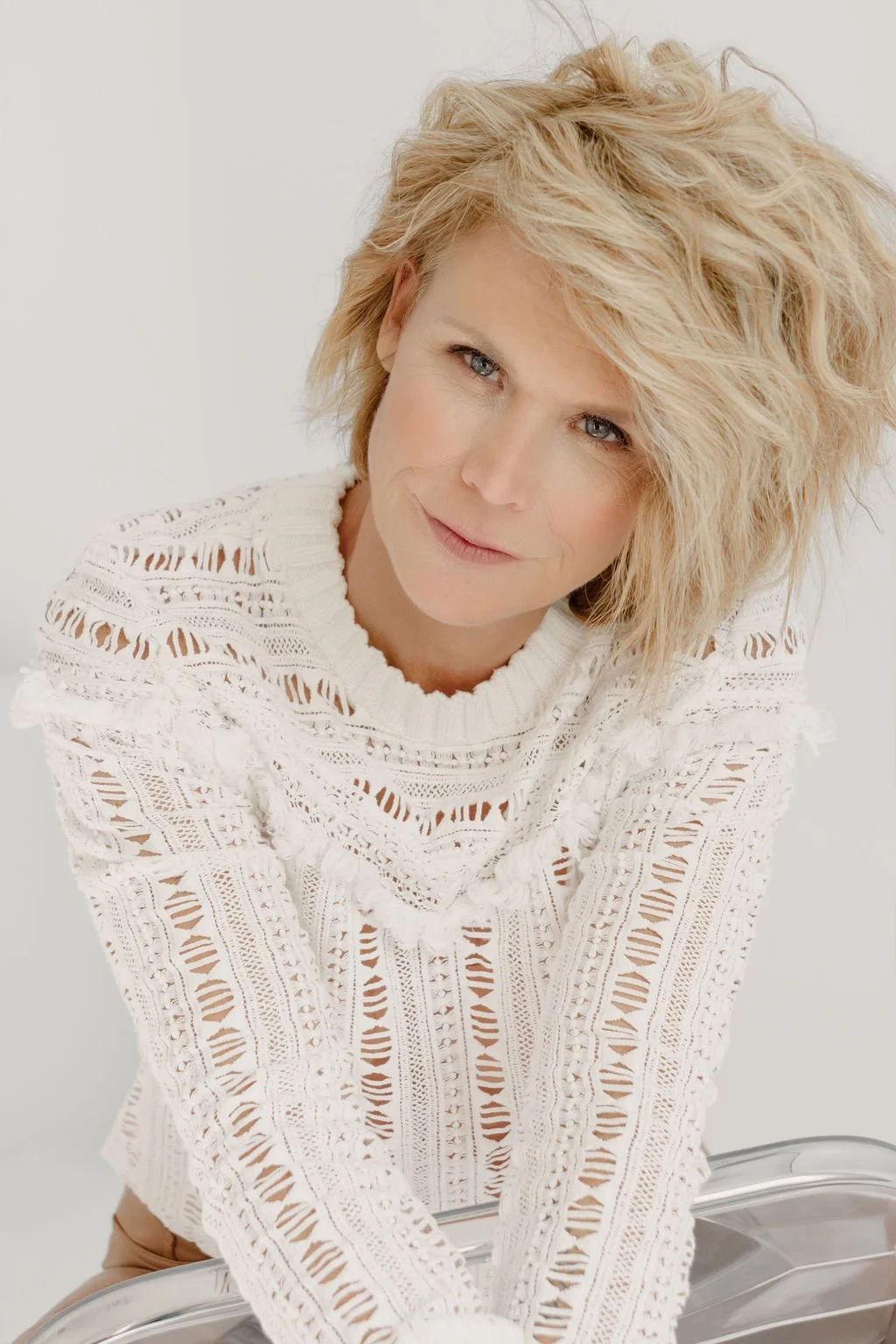 A woman with blonde, curly hair wearing a white, lacy, patterned sweater posing for a portrait against a plain background.