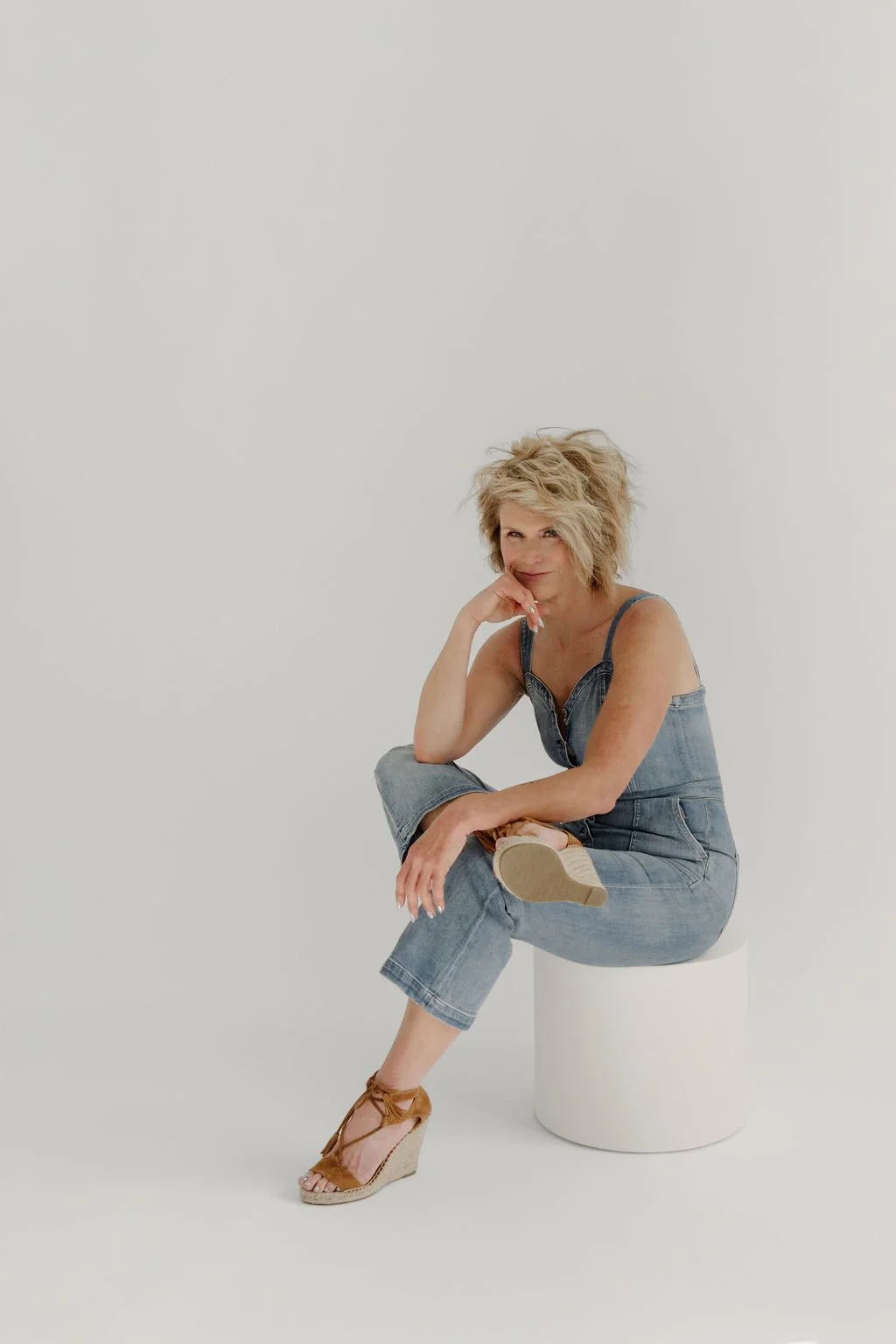 A woman sitting on a white cylindrical stool against a plain white background, wearing a denim jumpsuit and tan wedge sandals, with short, tousled blonde hair, looking at the camera with a playful smile.