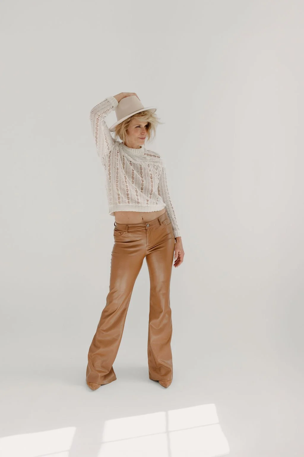 Woman in beige hat, loose crochet sweater, and flared tan leather pants standing in a white studio, holding the hat with one hand.