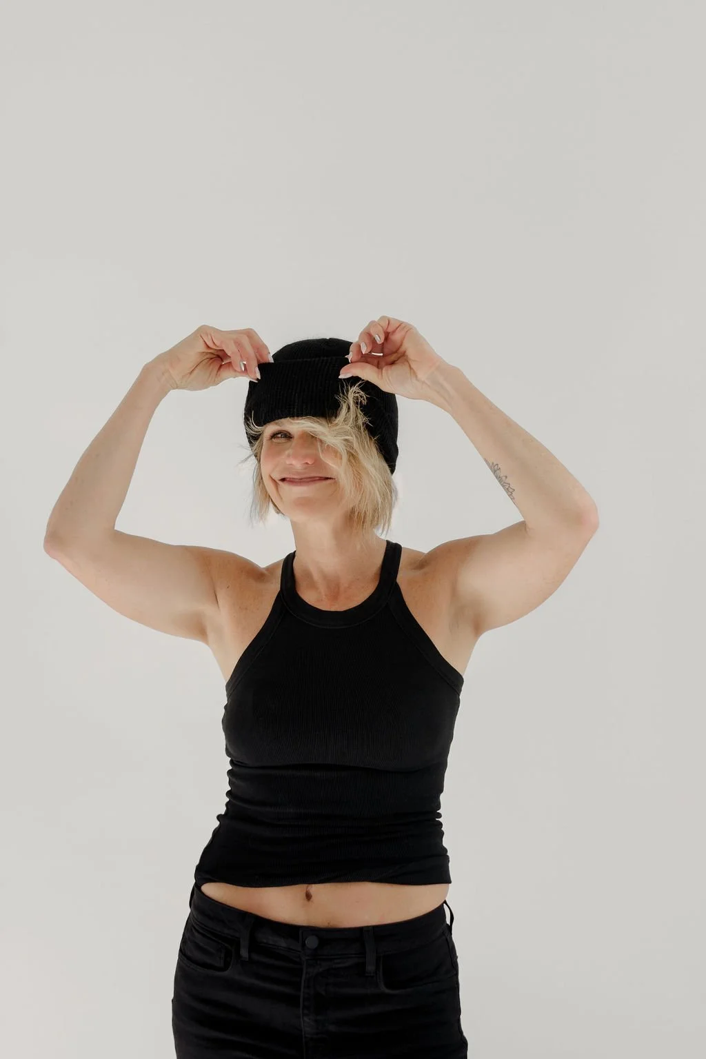 A smiling woman with blonde hair, wearing a black beanie, black tank top, and black pants, adjusting her beanie against a plain white background.