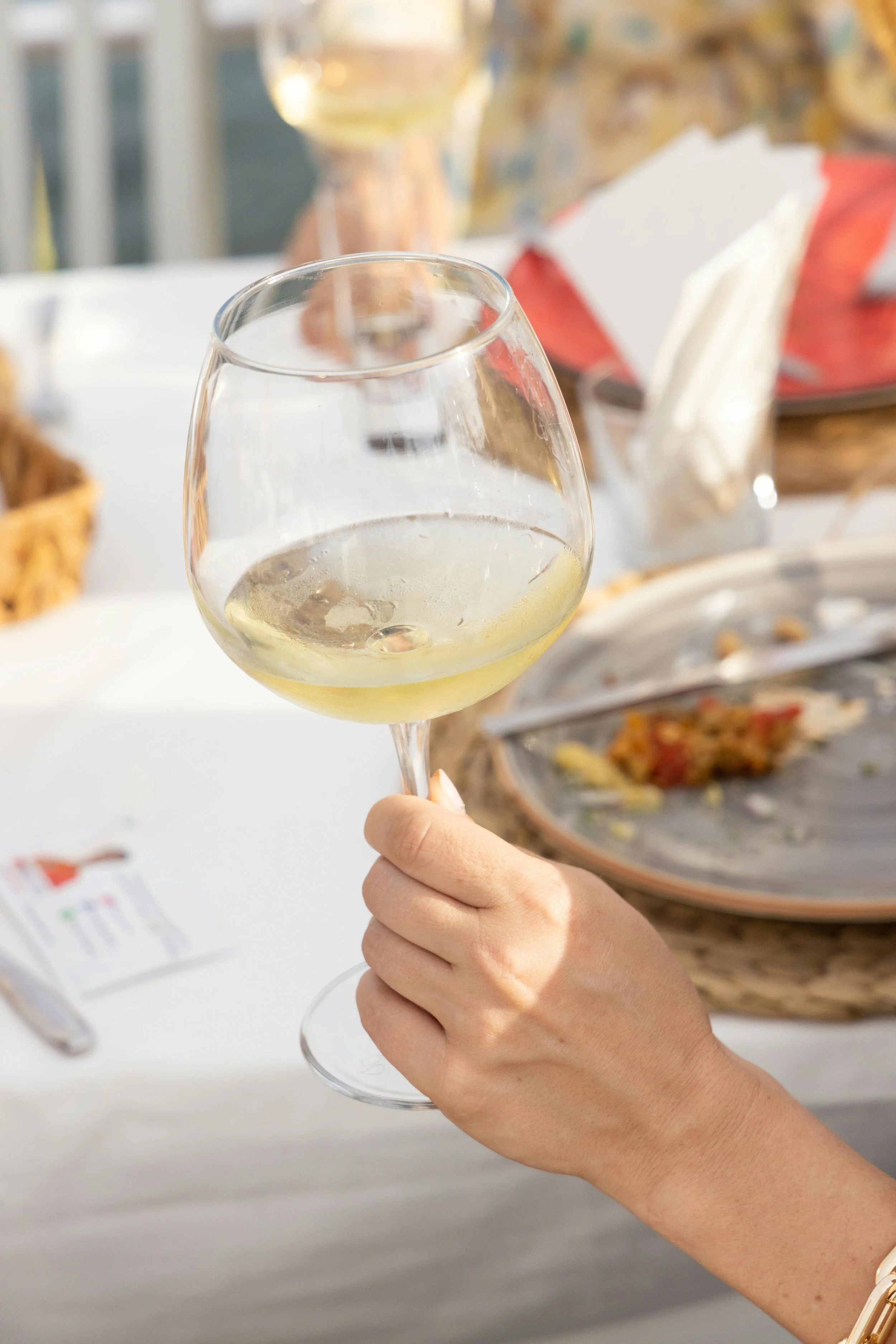 Persona sosteniendo una copa de vino blanco en una celebración. Fondo con comida y decoraciones.