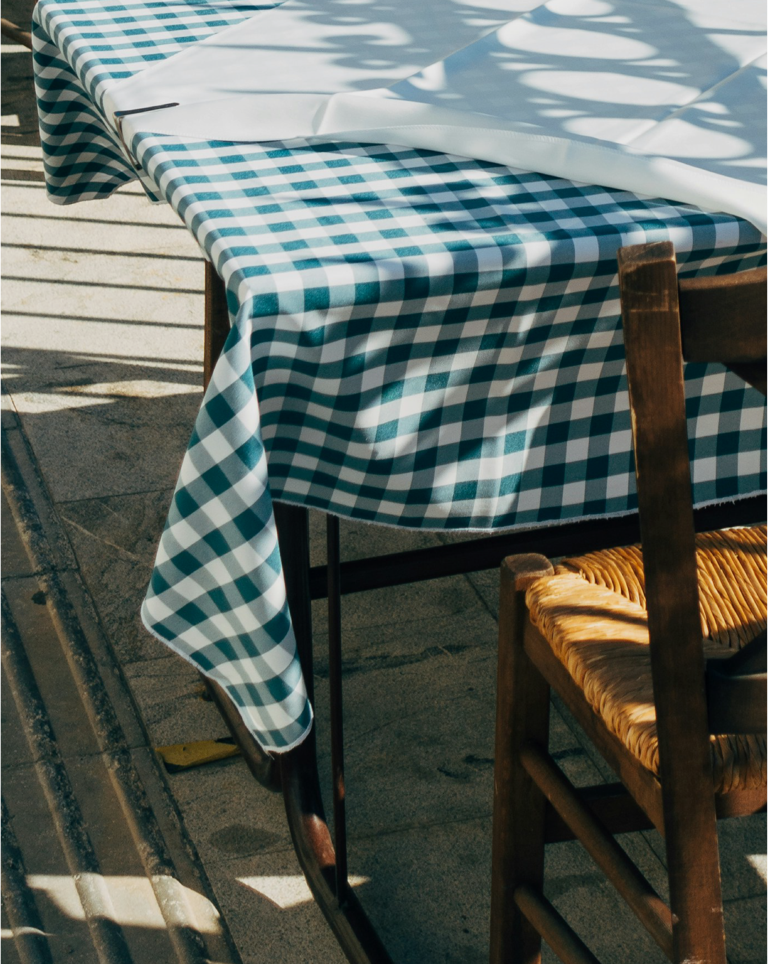 Mesa cubierta con tela de cuadros azul y blanco, con sombra de plantas sobre ella, y alrededor varios sillones de madera con asiento de ratán.