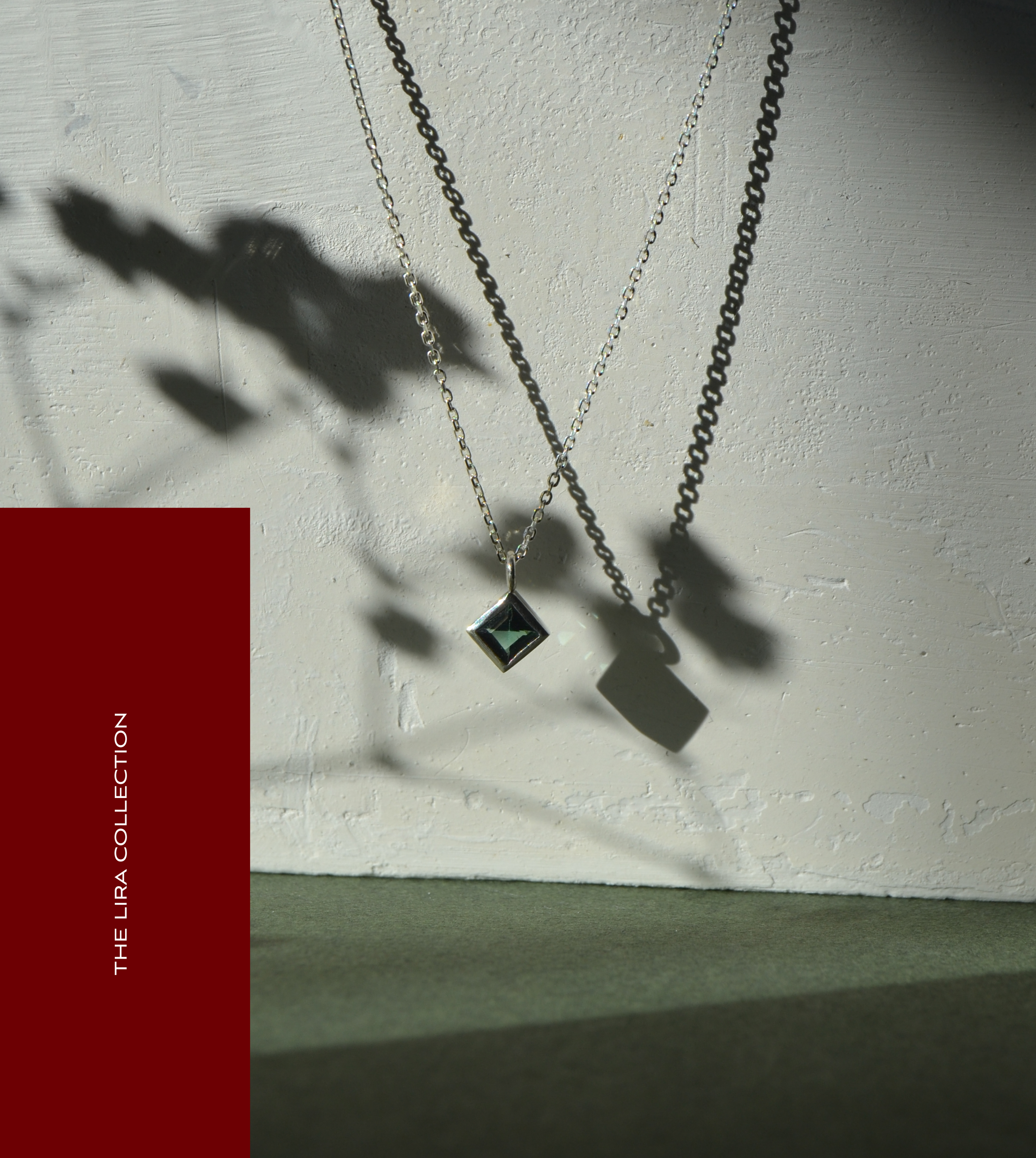 Joyería con cadenas de plata y un pendiente con gema verde colgado, proyectando sombras sobre una pared blanca y sobre una mesa de color gris oscuro.
