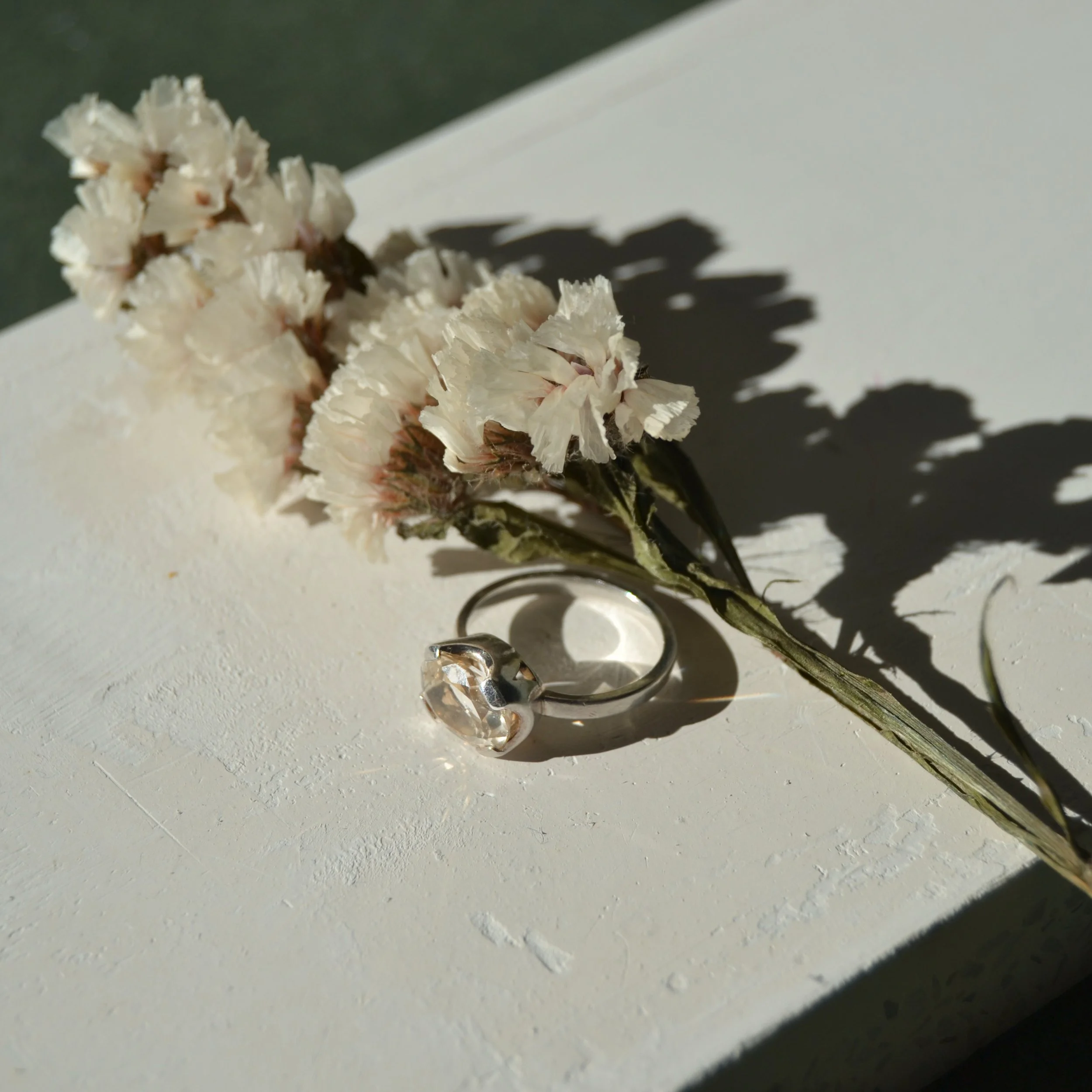 Ramos de flores blancas con un anillo de compromiso de diamantes junto a ellas, sobre una superficie blanca y con sombra