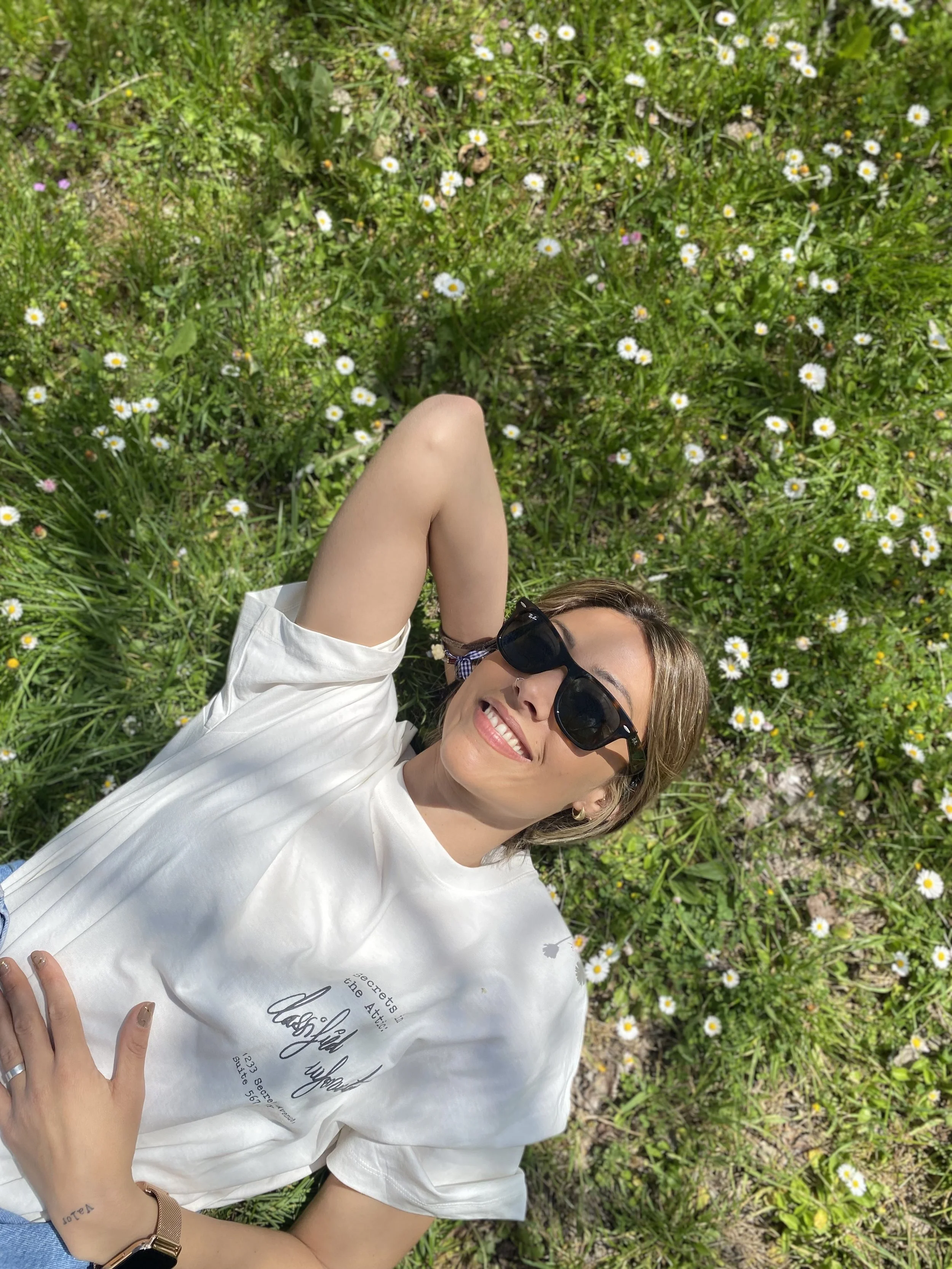 Mujer joven con gafas oscuras y blusa blanca acostada en la hierba con flores pequeñas y blancas con centro amarillo, descansando y sonriendo en un día soleado.