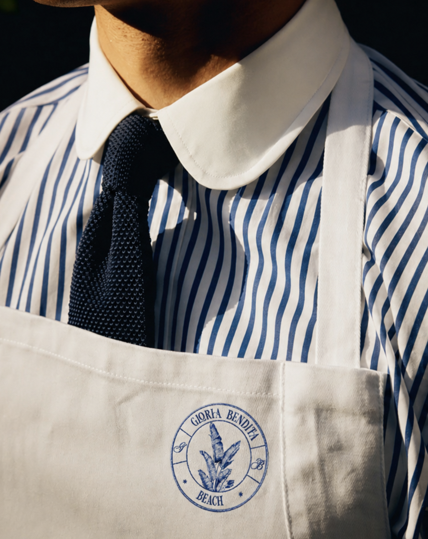 Persona con camisa a rayas y lazo negro, usando delantal con el logotipo de Gloria Bendita Beach.