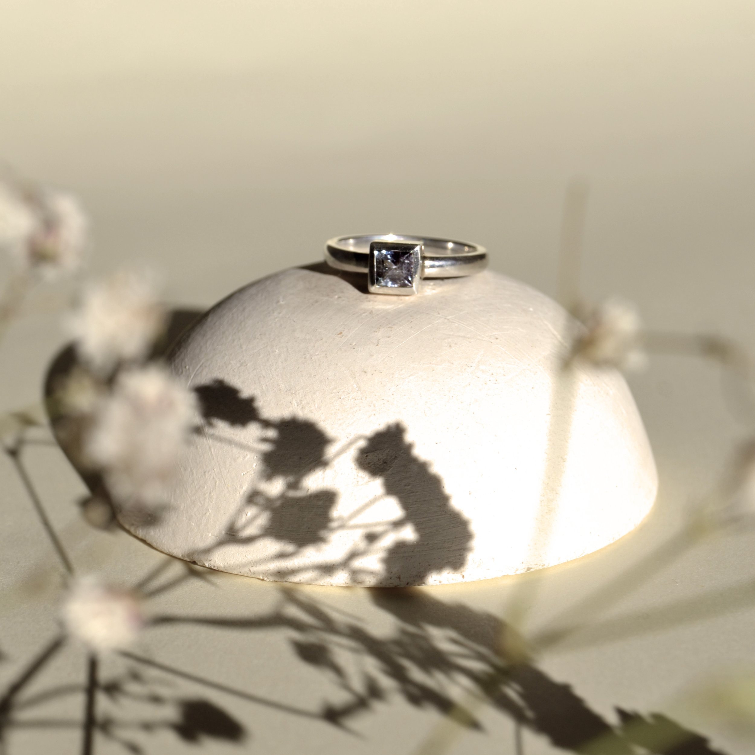 Un anillo de plata con una piedra cuadrada en el centro, colocado sobre una superficie que parece una piedra. Se proyectan sombras de ramas secas en la superficie.