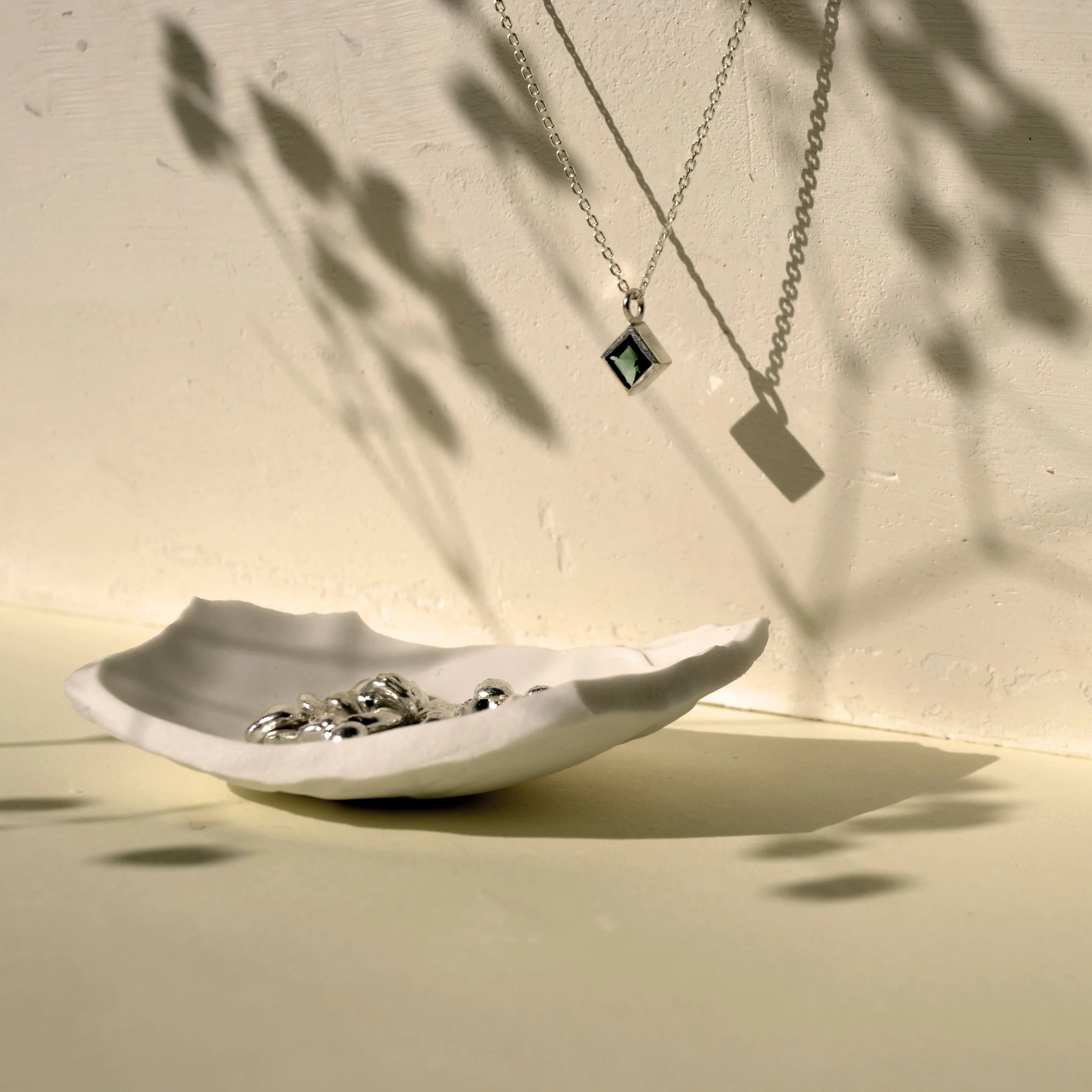 Collar de plata con colgante rectangular que tiene piedra verde, colgado en una pared beige con sombra de hojas y una escena de una pieza en una olla plateada en la parte inferior.