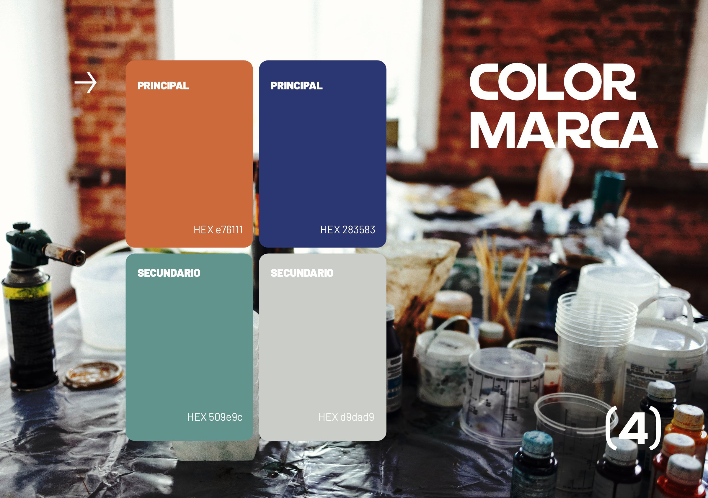 Mesa de trabajo con materiales de pintura y arte, como botellas de pintura, pinceles y recipientes, en un espacio con paredes de ladrillo y ventanas