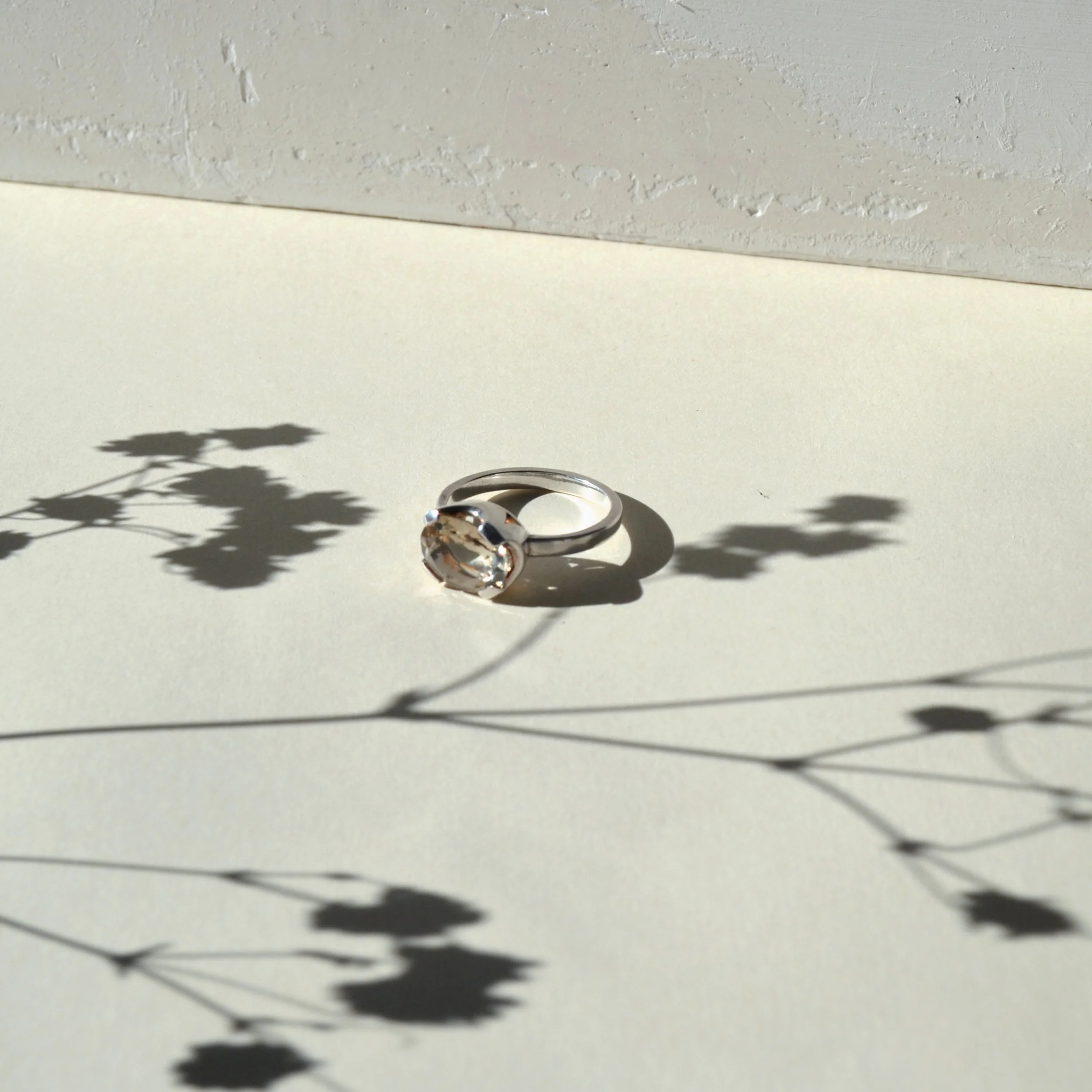 Un anillo de plata con una piedra brillante sobre una superficie blanca, proyectando sombras y reflejos.