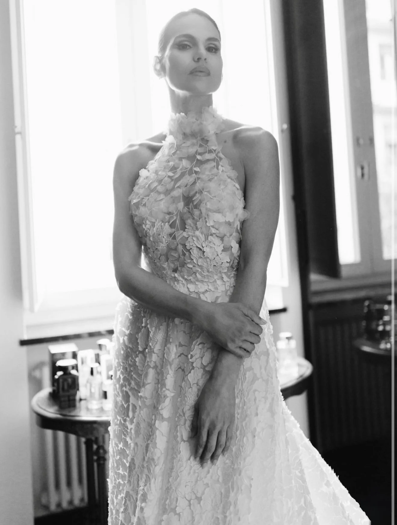 Una donna in abito da sposa elegante, senza braccio, scattato in bianco e nero, in piedi in una stanza con finestra luminosa sullo sfondo.