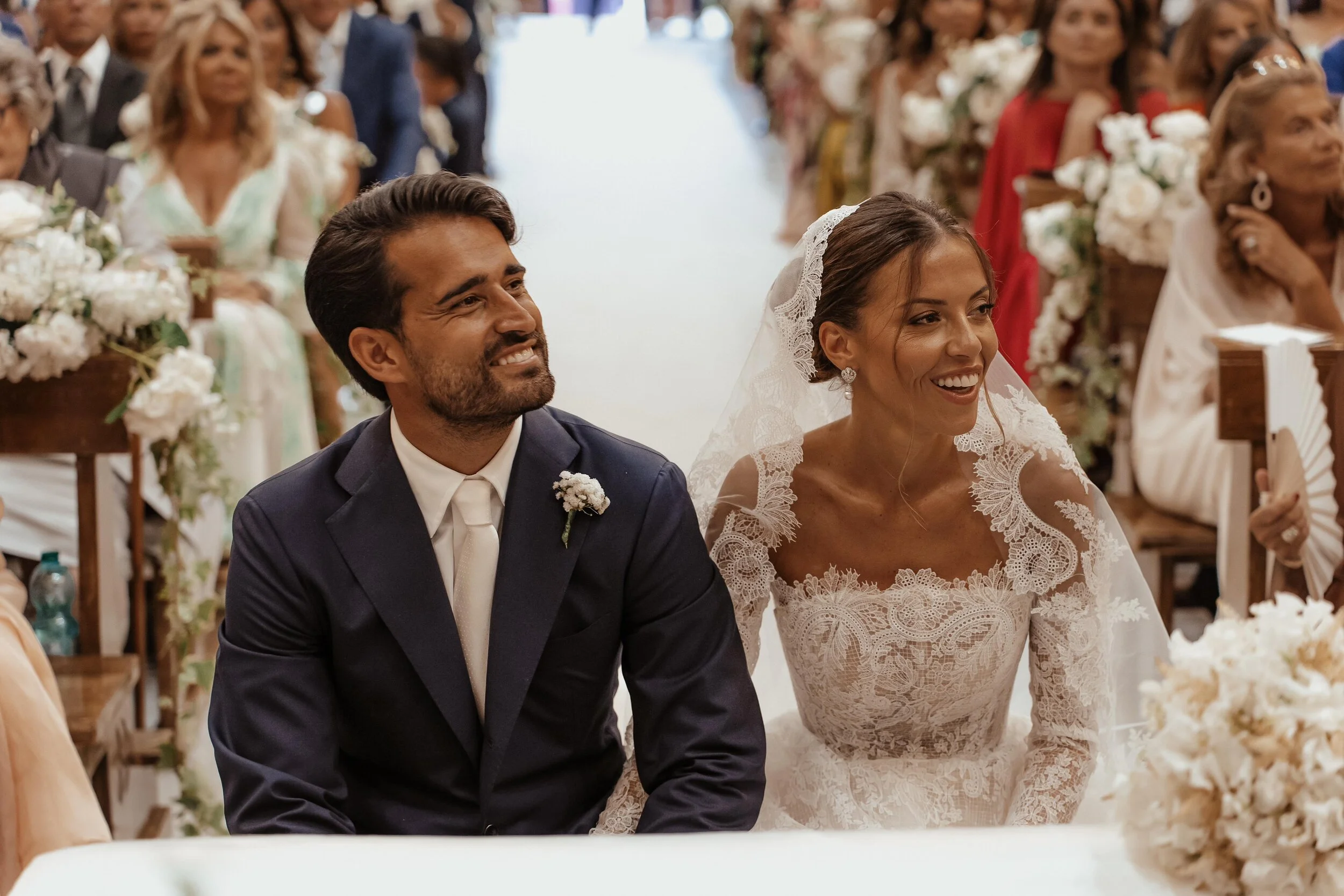 Nozze in chiesa con sposi seduti, donna in abito da sposa con velo e uomo in abito scuro, presenti invitati seduti con bouquet di fiori.