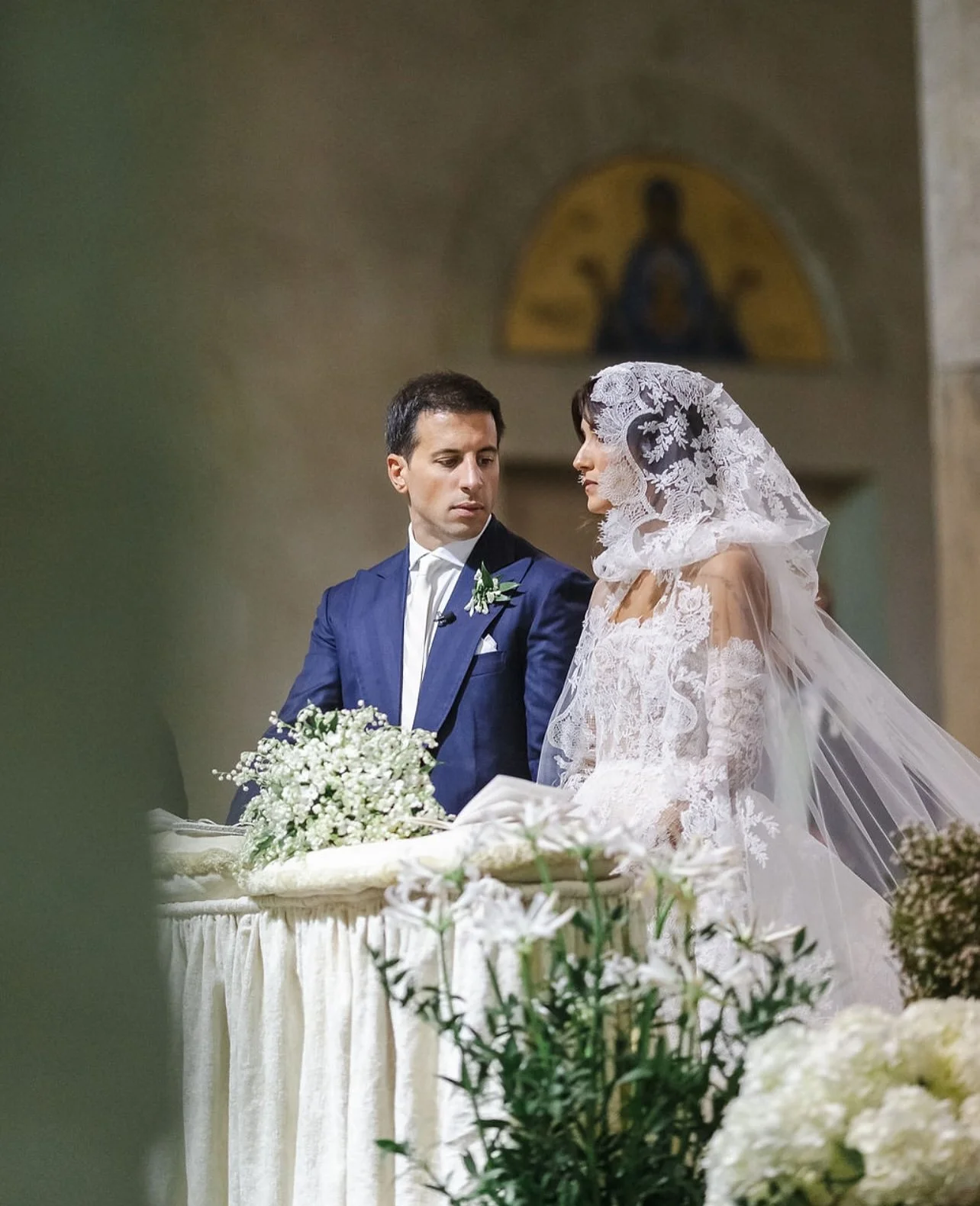 Una coppia di sposi durante il matrimonio, con il marito in abito blu scuro e la sposa in un abito da sposa bianco con velo e mantiglia ricamata, si trovano davanti a un altare decorato con fiori bianchi, in una chiesa con affresco alle spalle.