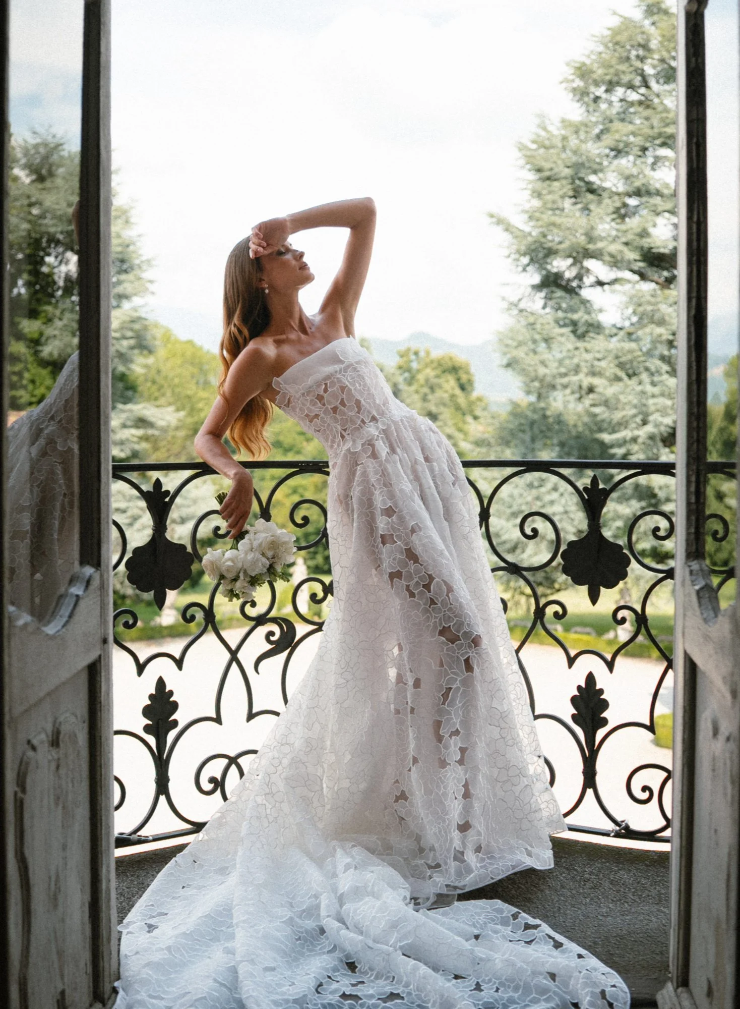 Una donna in un abito da sposa bianco con ricami, che si staglia su un balcone con ritratto di paesaggio naturale con alberi e cielo.