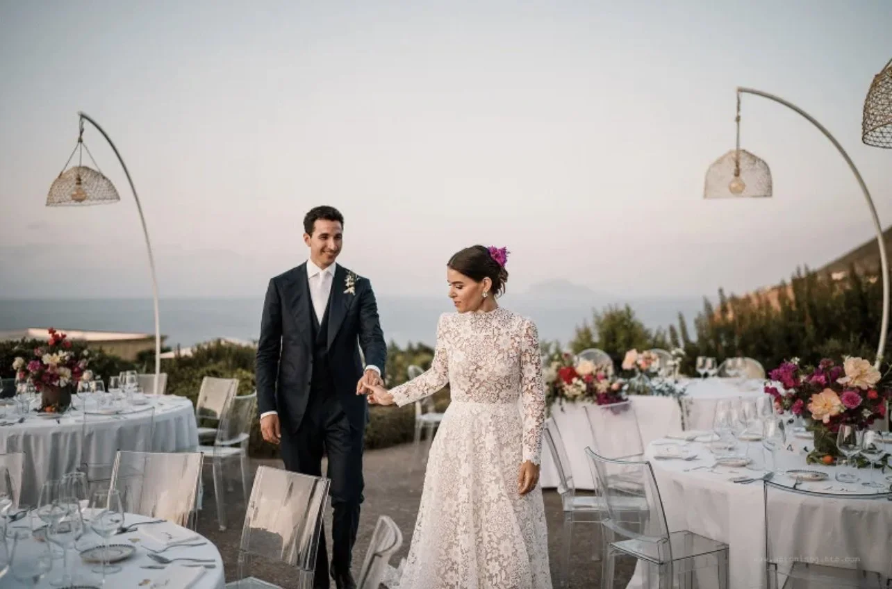 Sposi in abiti da matrimonio camminano mano nella mano durante un ricevimento all'aperto al tramonto, con tavoli decorati con fiori e bicchieri, sullo sfondo vista sul mare e colline.