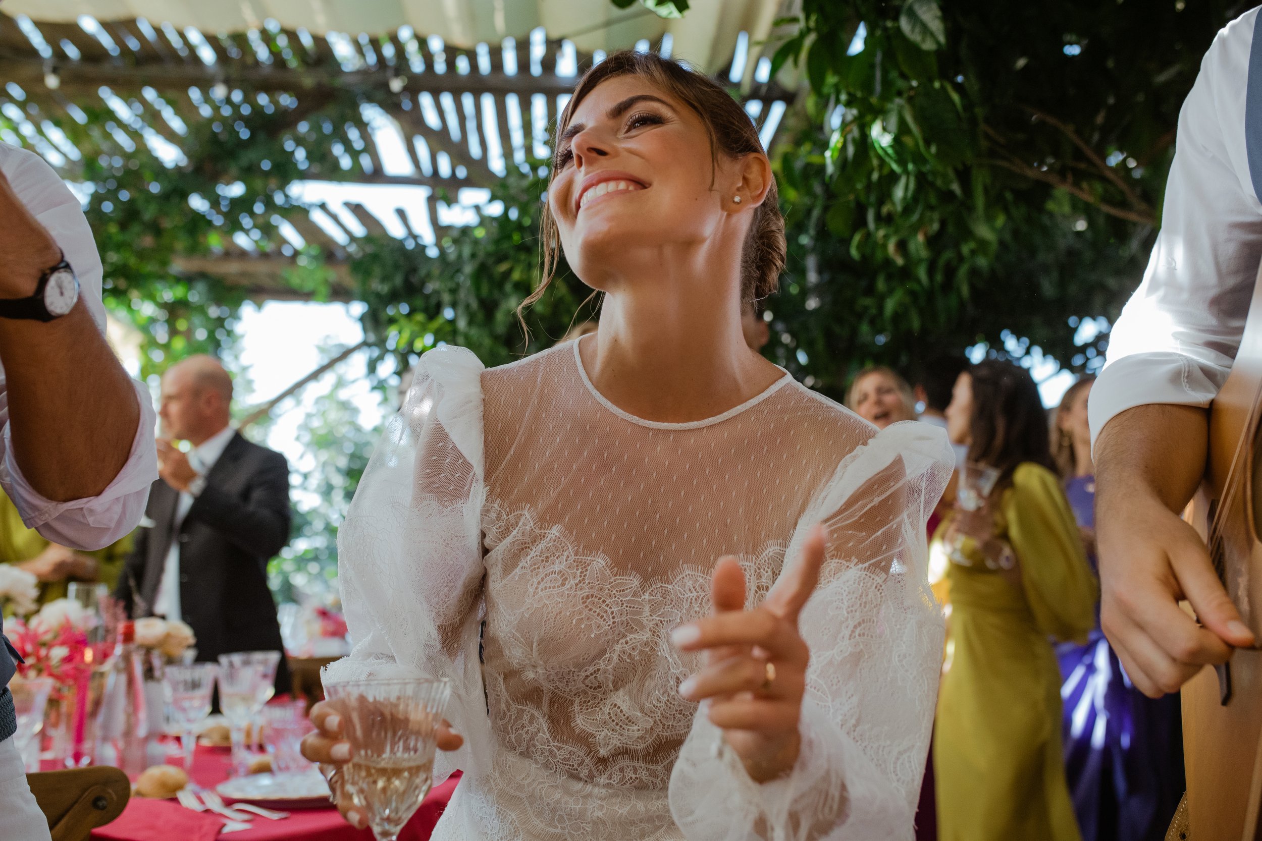 Donna sorridente con abito bianco in un evento all'aperto, circondata da altre persone, alcune con abiti colorati, sotto un gazebo di legno con luci naturali e vegetazione.