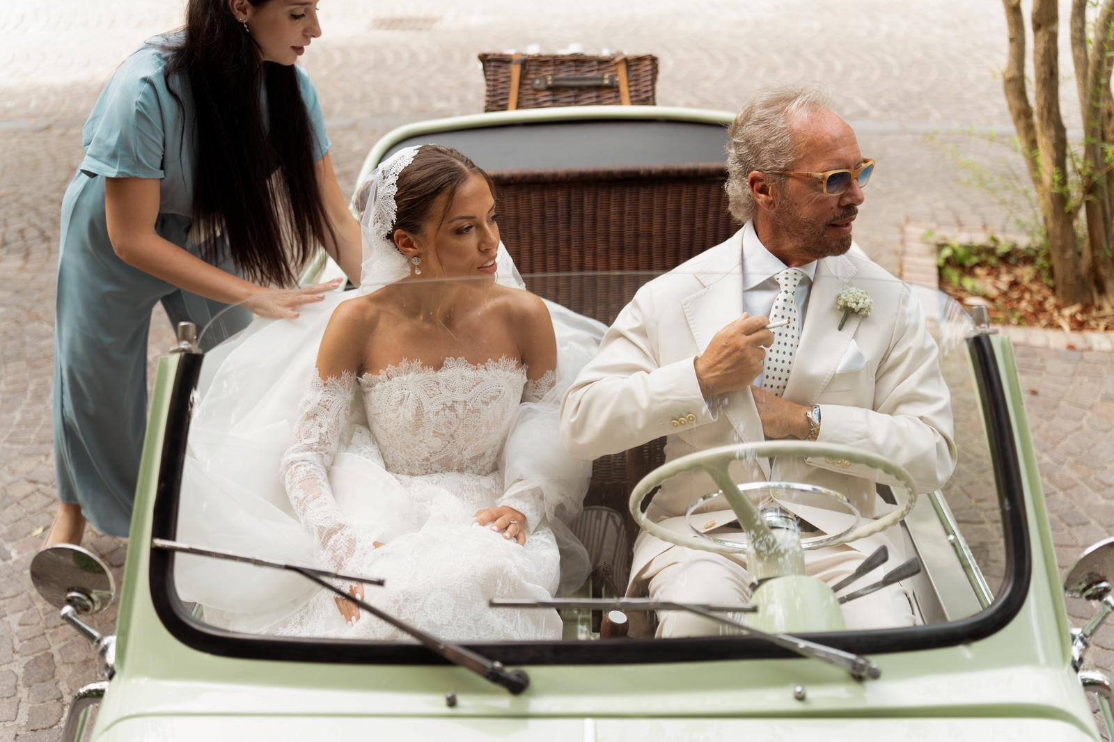 Sposi in auto d'epoca, la sposa seduta e il padre al volante, assistita da una donna dietro, all'aperto in un'area pavimentata.