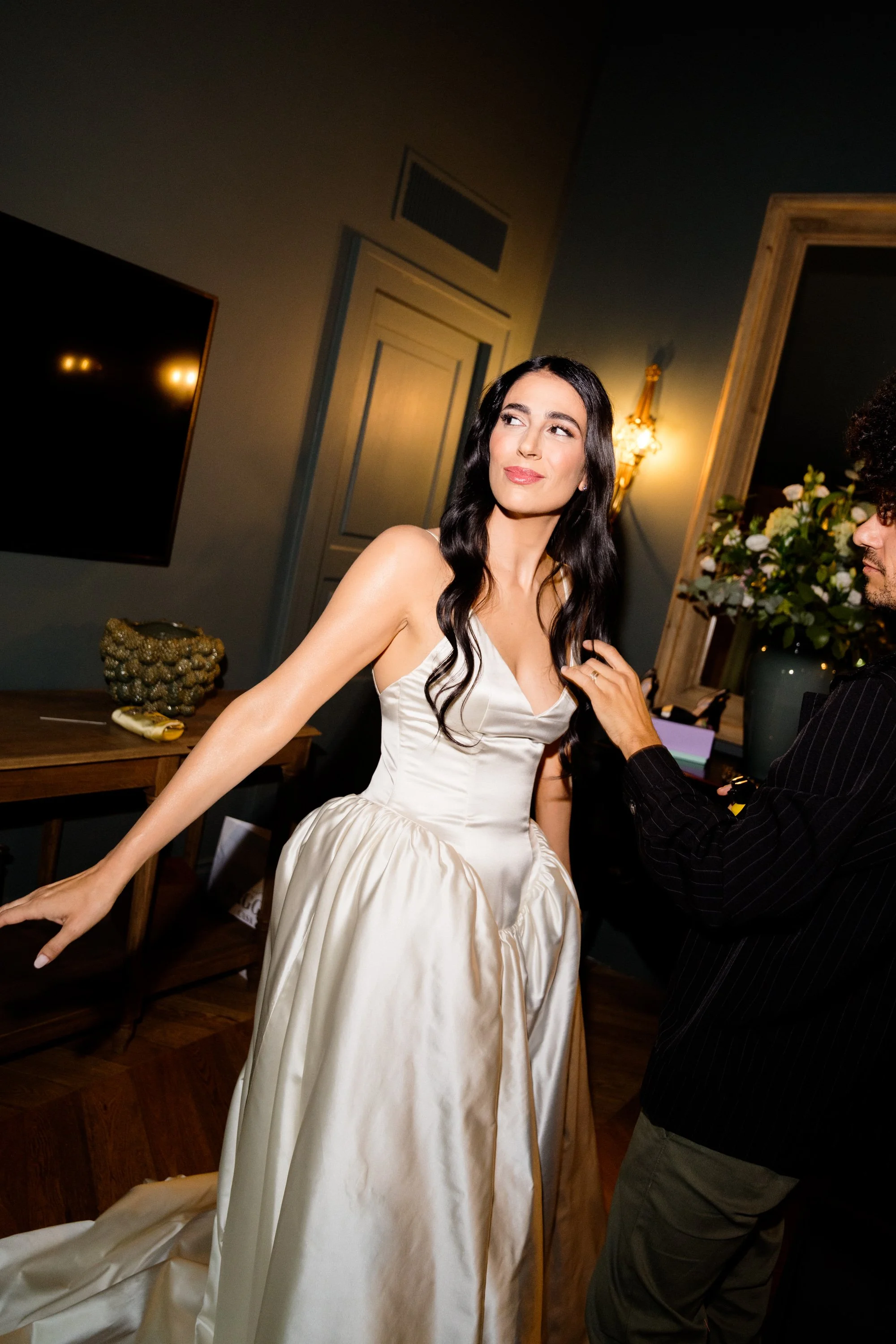 Una donna che indossa un abito da sposa in attesa di essere sistemata, mentre un assistente le sistema i capelli in un ambiente elegante.