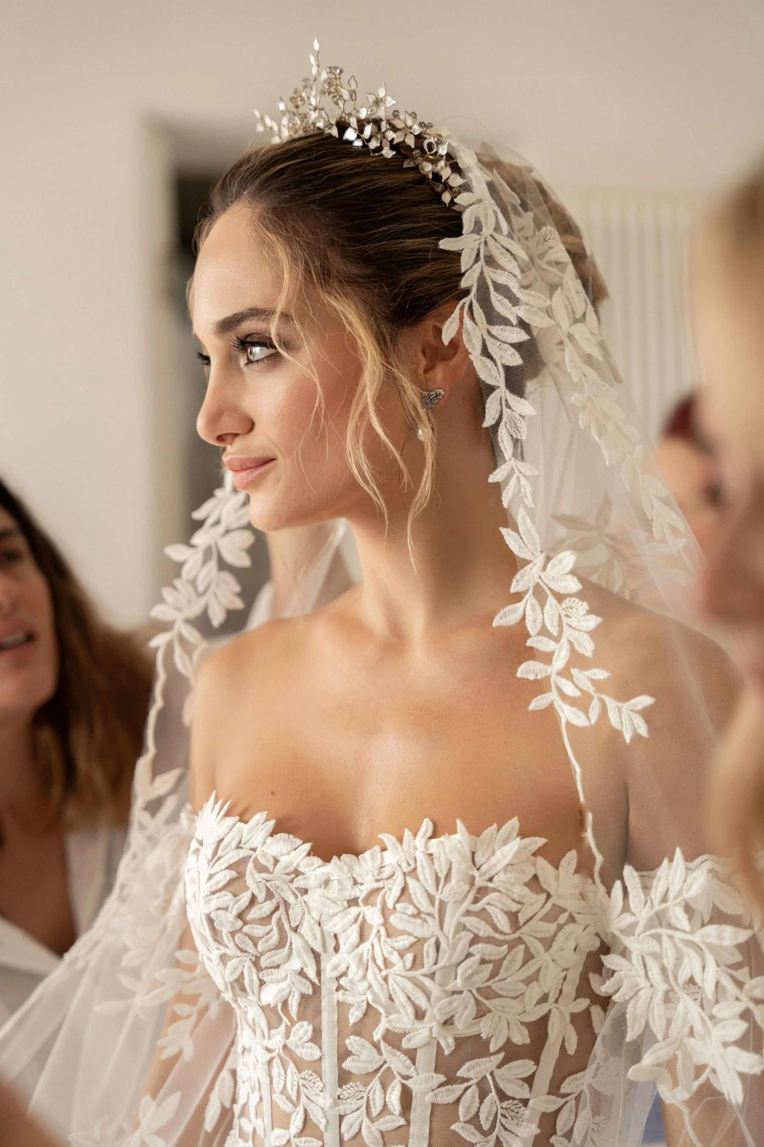 Una sposa che indossa un abito da sposa con dettagli in pizzo e una tiara, con un velo decorato con foglie di pizzo, in un ambiente interno.
