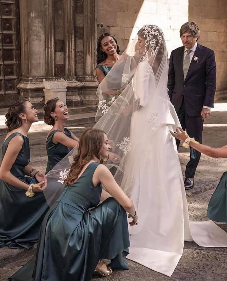 Servizio fotografico di nozze con cinque donne e due uomini in un luogo storico. La sposa indossa un abito bianco con velo e laurea, circondata da amiche in abiti blu, mentre un uomo in abito scuro rivolge uno sguardo alla scena.