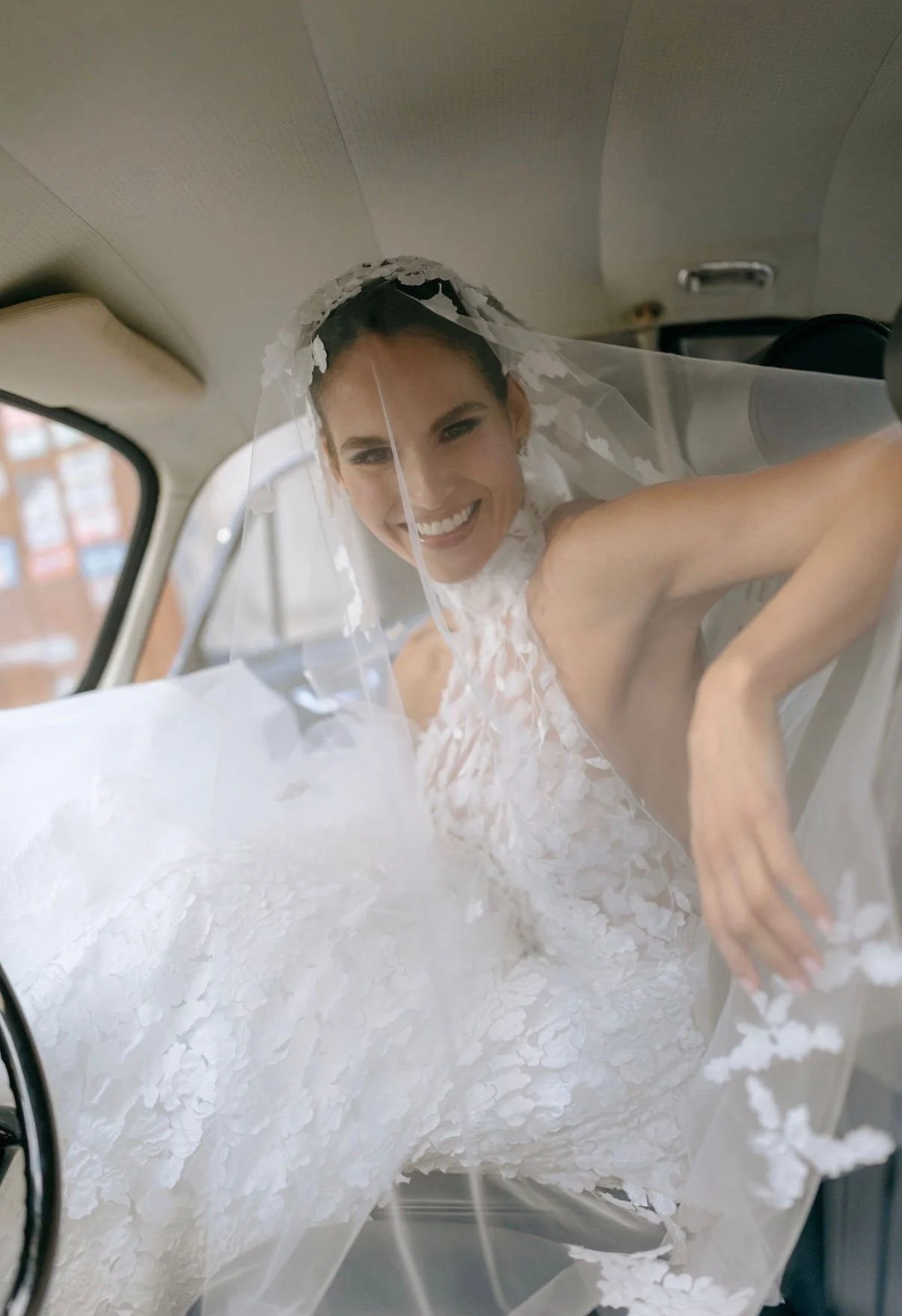 Sposa in abito da matrimonio seduta nell'auto, coperta da velo e sorridente.