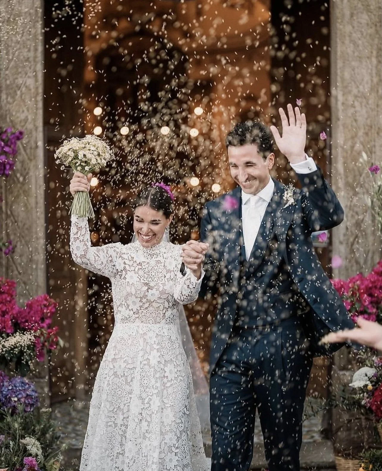 Una coppia di sposi felici che si festeggiano con confetti sotto un arco decorato di fiori durante il matrimonio.