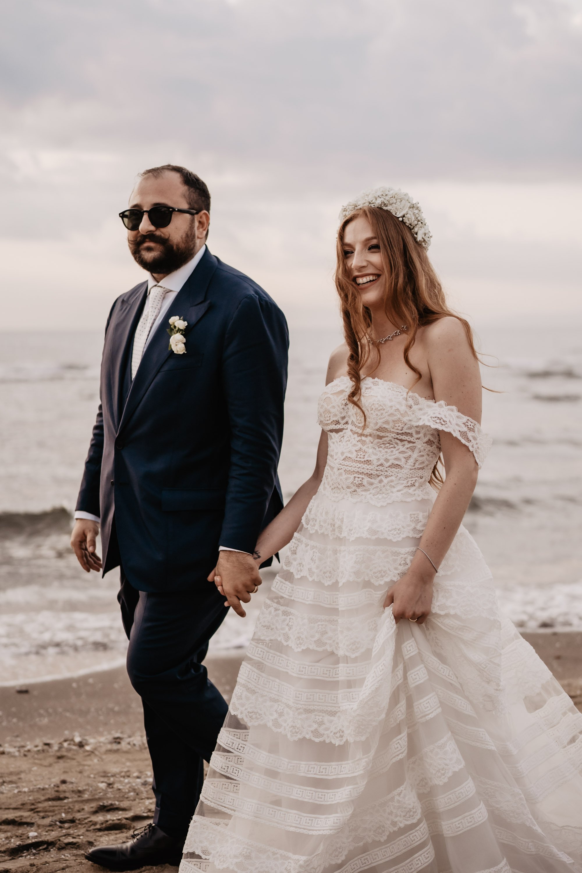 Coppia di sposi sulla spiaggia, la donna indossa un abito da sposa bianco con dettagli in pizzo, lui un abito blu scuro con camicia bianca e cravatta, si tengono per mano