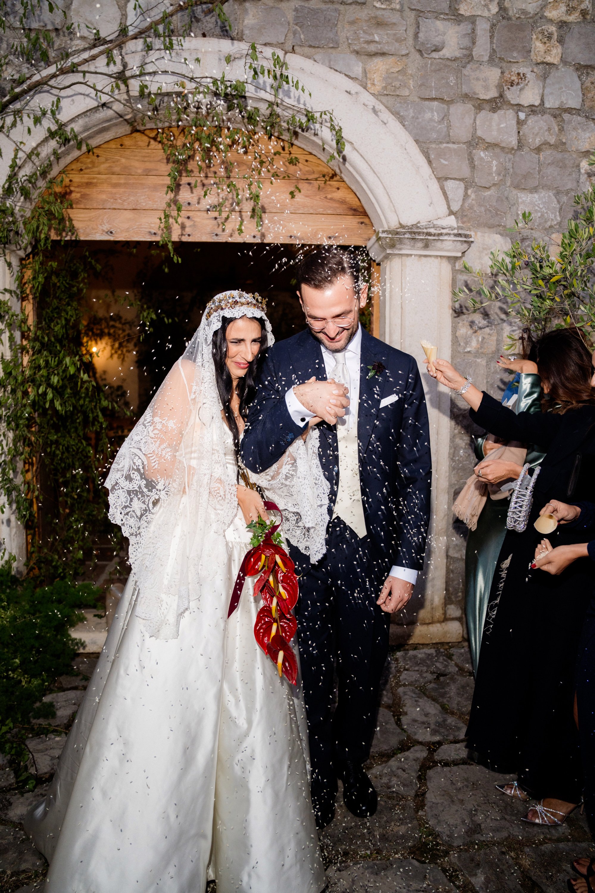 Sposi di matrimonio in abito tradizionale, la sposa tiene un bouquet rosso, vengono spruzzati con confetti da amici all'ingresso di un edificio in pietra.