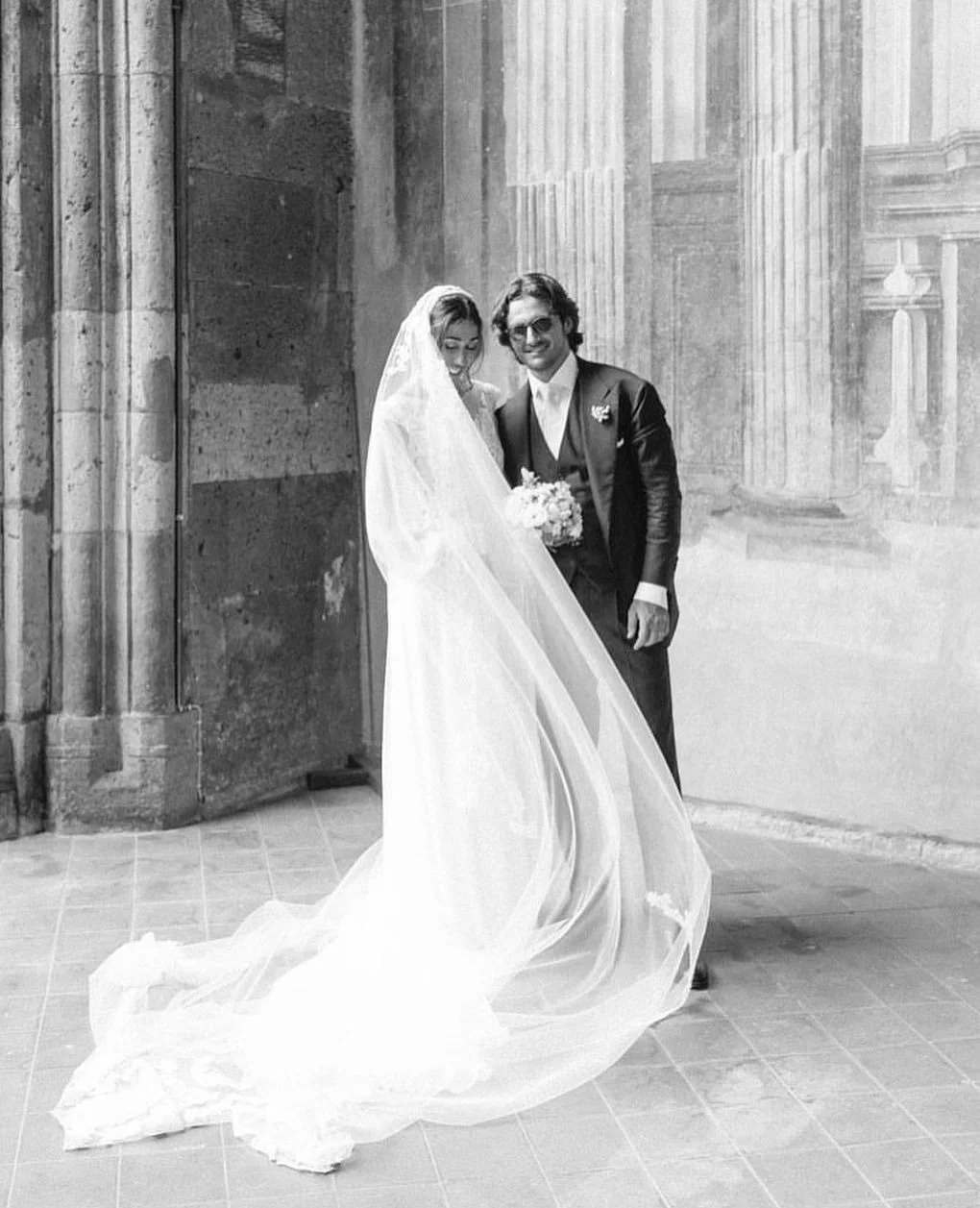 Sposo e sposa sorridenti in abiti da matrimonio, con sfondo di colonne antiche e architettura storica.