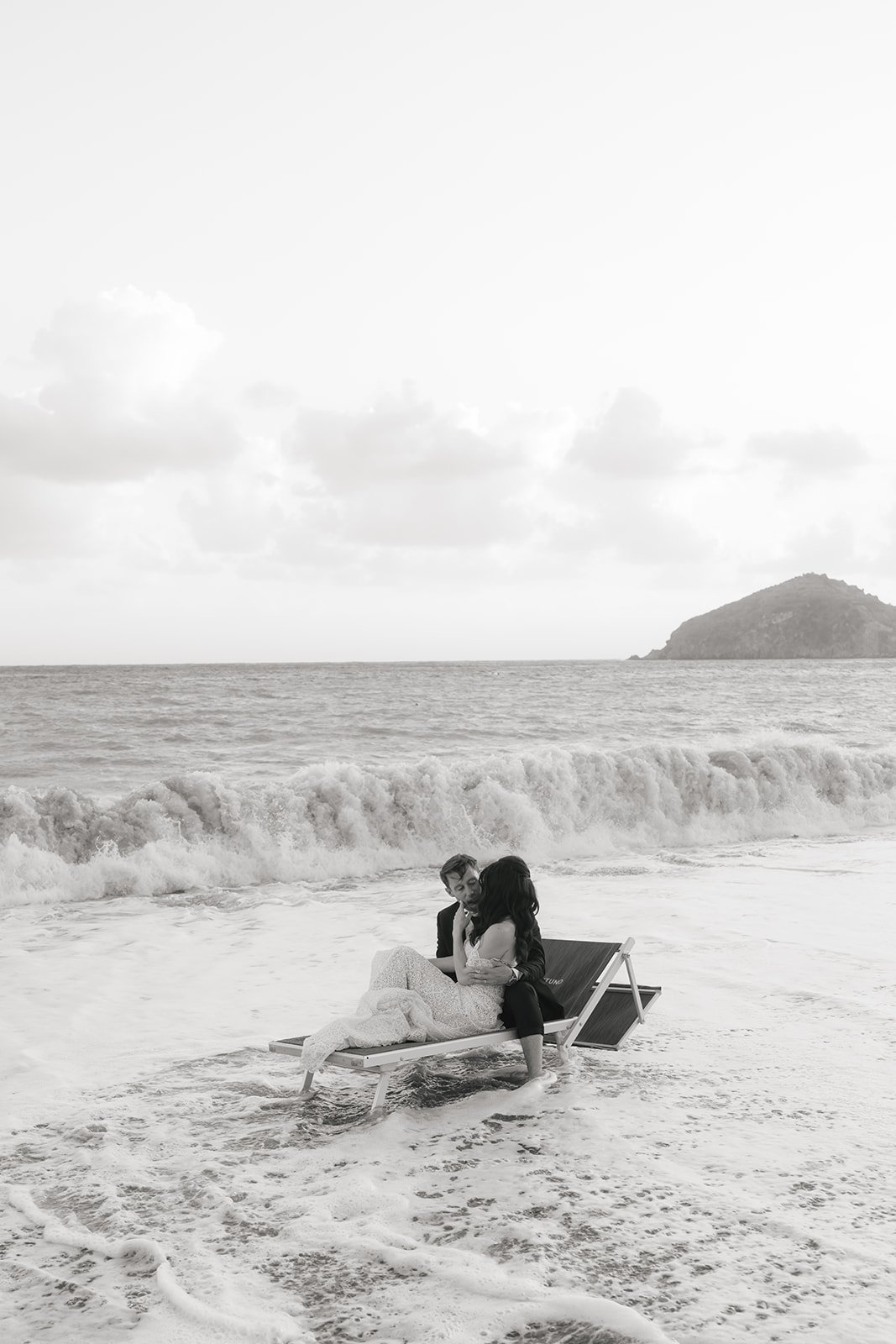 Due persone, una donna in un abito bianco e un uomo in abito scuro, sedute su una sdraio sulla spiaggia vicino all'acqua.
