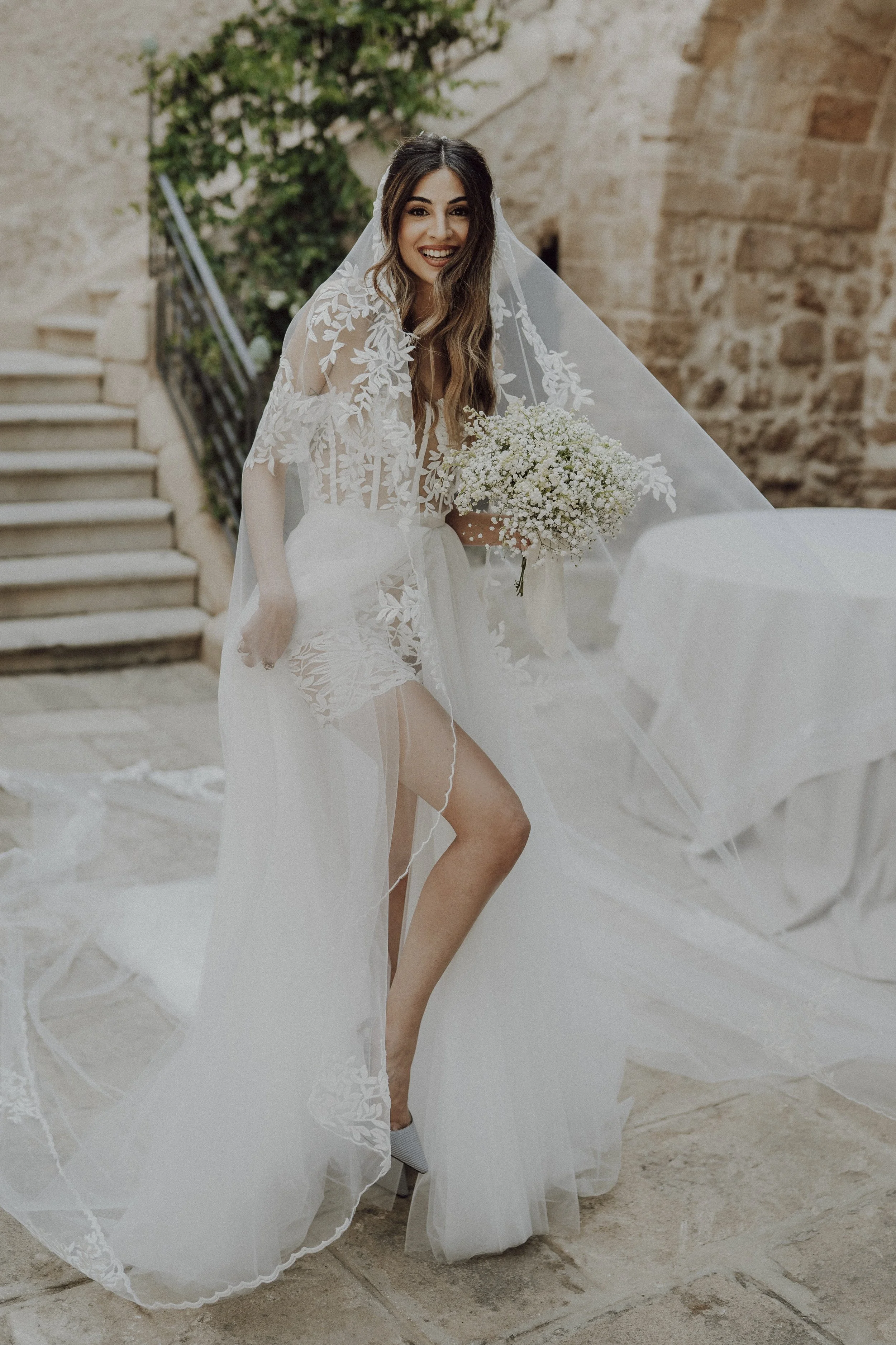 Una donna in abito da sposa con un velo ricamato sorride, tiene un bouquet di fiori bianchi, circondata da un ambiente antico di pietra e scale.