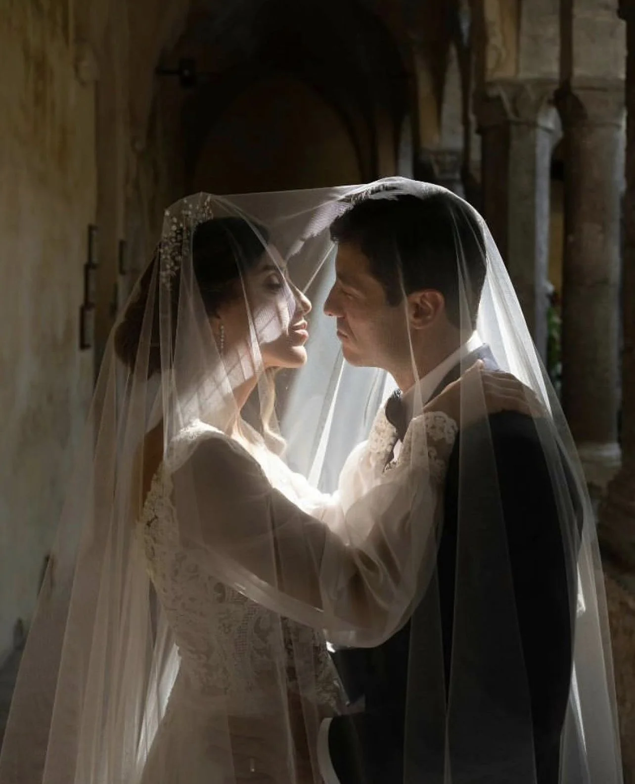 Una coppia di sposi si bacia sotto un velo da sposa in una location storica con archi.