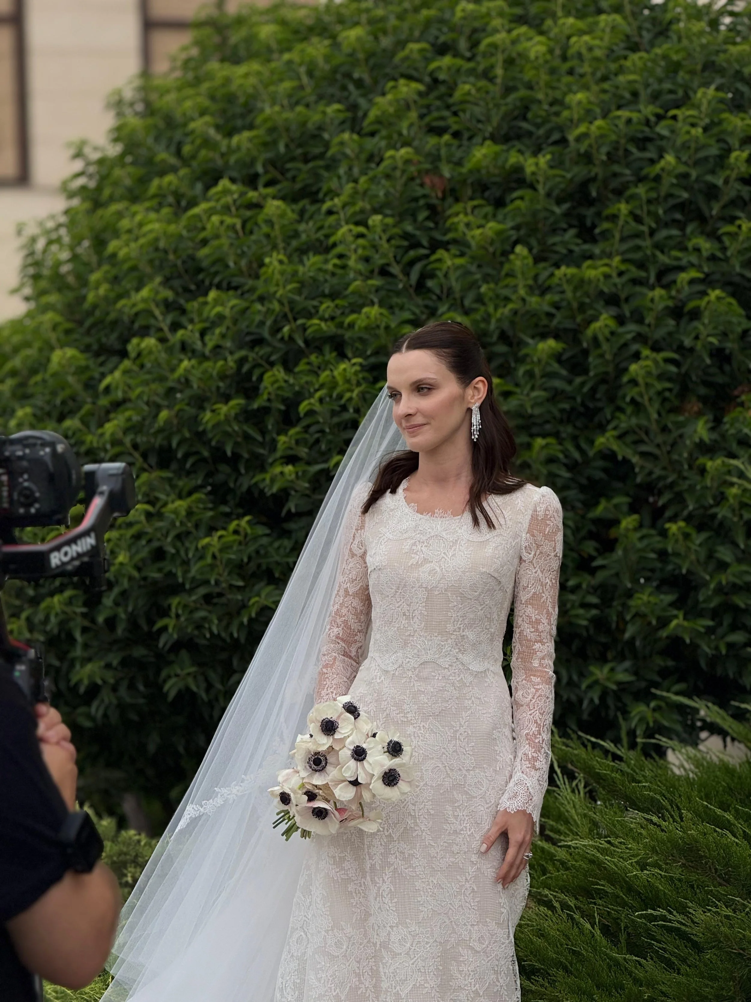 Una sposa con abito da sposa in pizzo e veli, che tiene un bouquet di fiori bianchi con centri neri, si trova all'aperto davanti a un cespuglio verde, mentre un fotografo la sta scattando.
