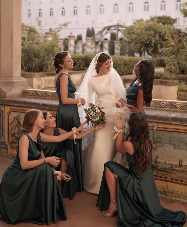Una coppia di sposi con le loro amiche sorridenti, che si scambiano regali e abbracci, durante la cerimonia di un matrimonio in un luogo all'aperto con archi e vegetazione.