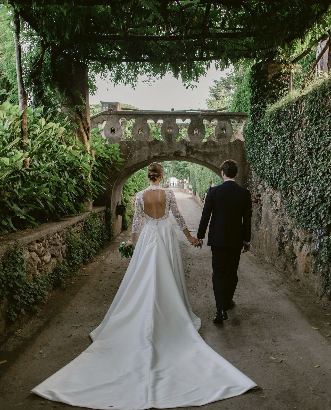 Coppia di sposi che cammina mano nella mano sotto un arco in un giardino, con la sposa in abito bianco e il velo lungo, e lo sposo in abito nero.
