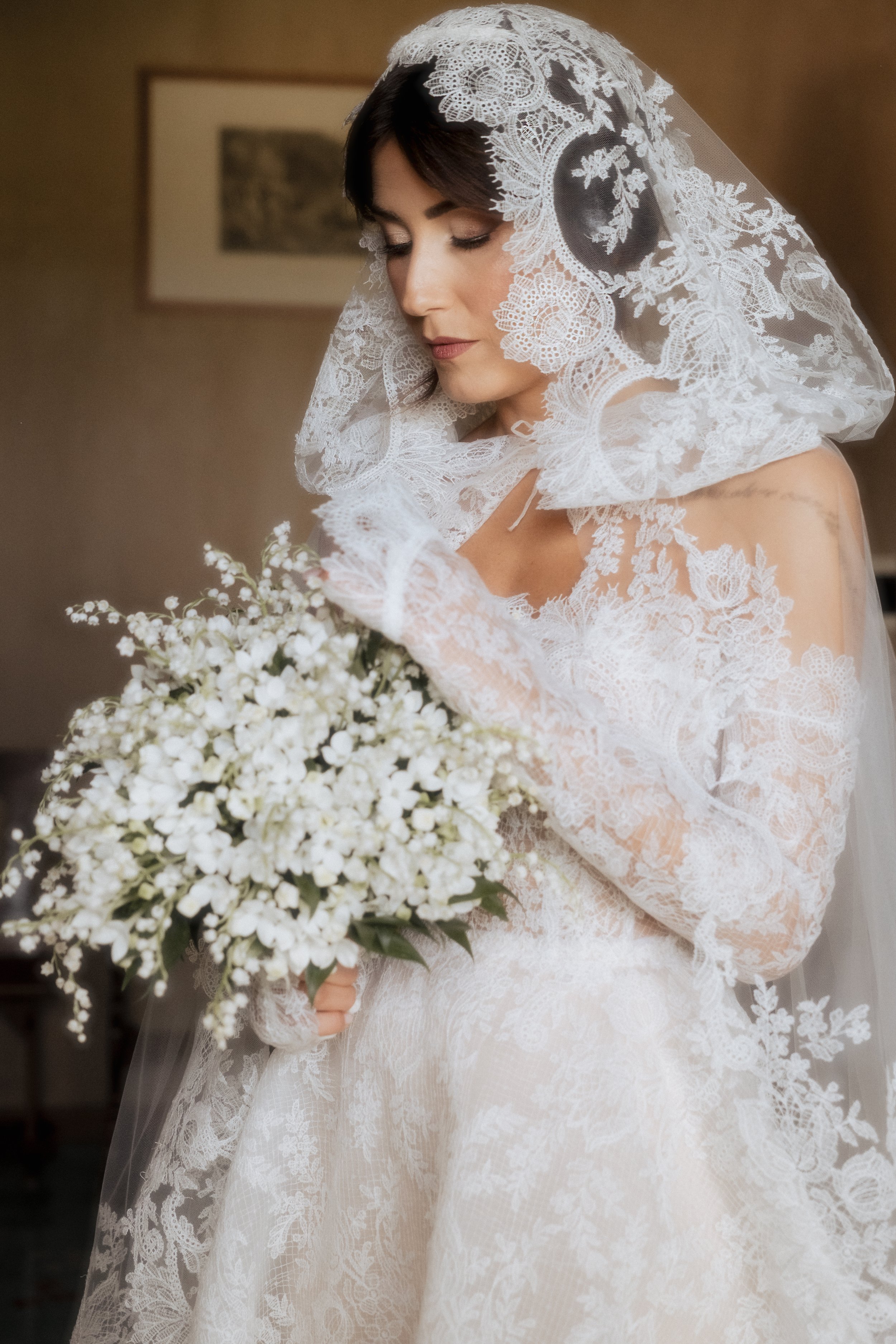 Una sposa con abito di pizzo bianco, in posa frontale, che tiene un bouquet di fiori bianchi, con un velo di pizzo chele copre leggermente il volto, in un ambiente interno con quadro sul muro.