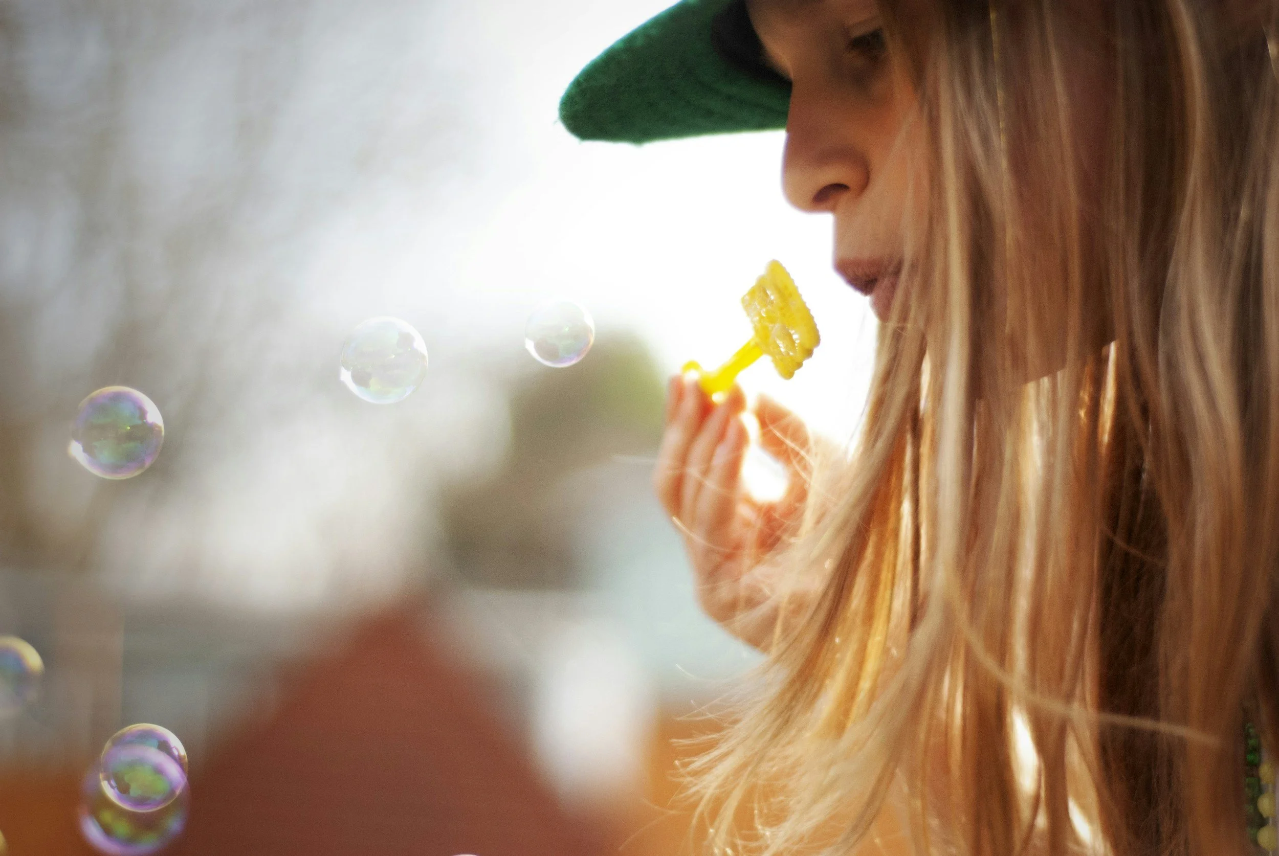 A woman with long, blonde hair blowing bubbles outdoors during daytime. She is wearing a green hat and appears to be enjoying the moment.