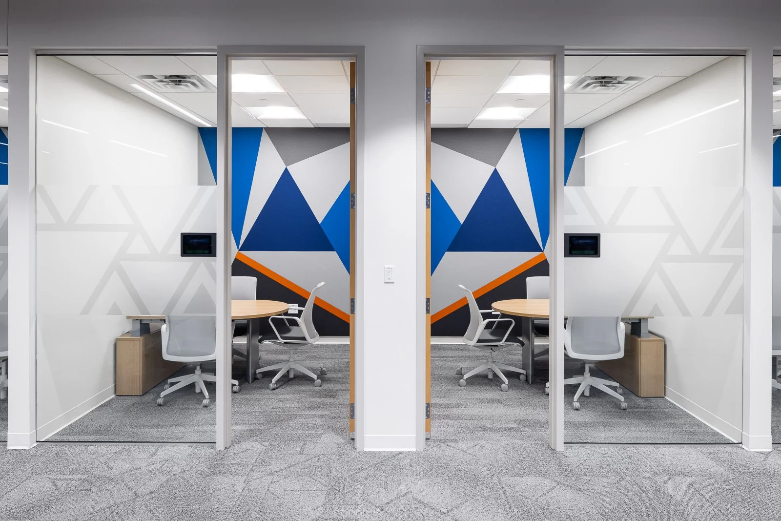 Two private office spaces with glass walls, featuring modern white chairs, round wooden tables, and a colorful geometric mural on the back wall with blue, gray, black, and orange shapes.