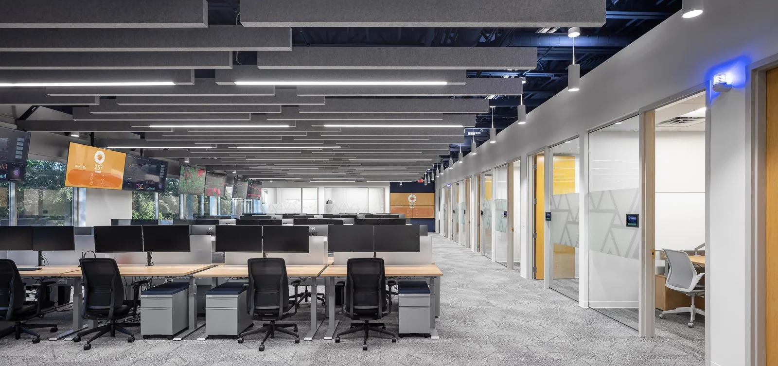 Modern office space with multiple workstations, black rolling chairs, and desk monitors. Glass office rooms with sliding doors line the right side, and digital displays and windows are visible on the left. The ceiling has gray acoustic panels and suspended lighting.