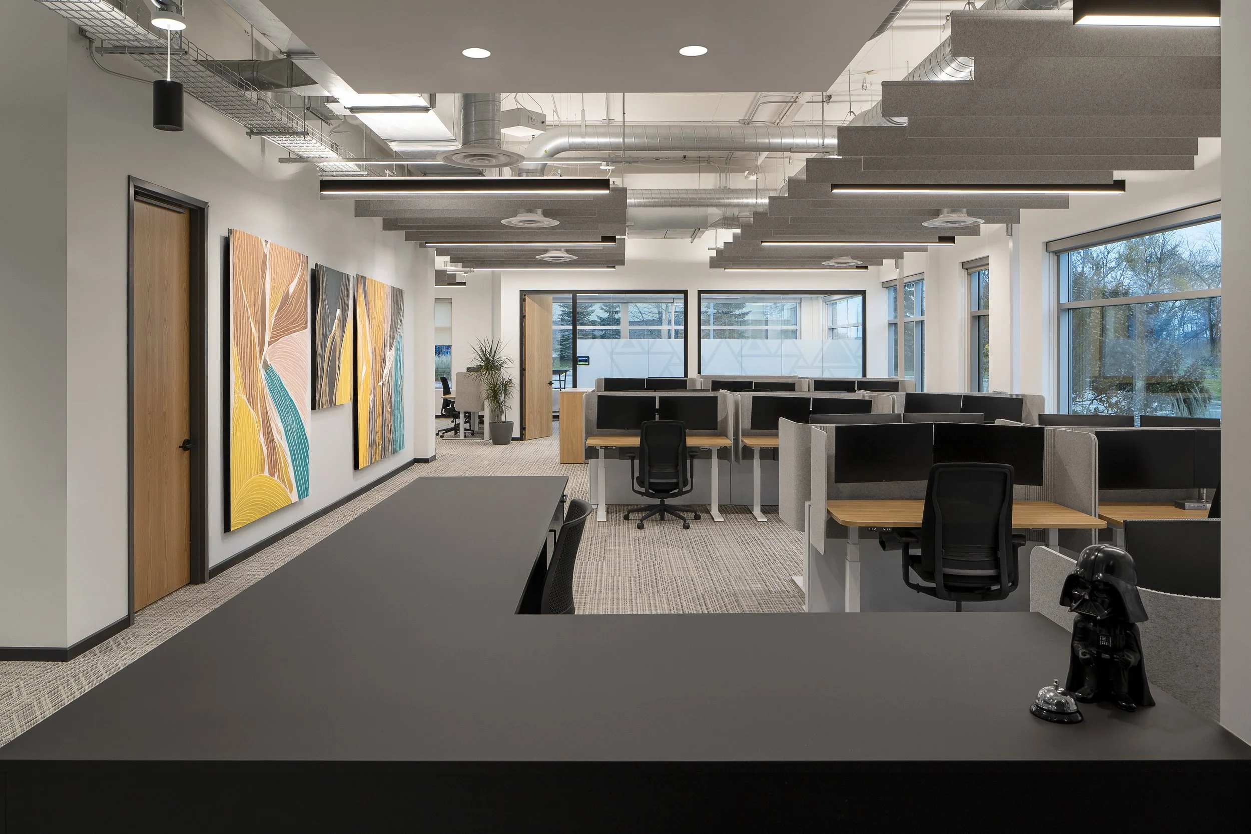 Modern office space with desks, chairs, large windows, wall art, and exposed ceiling pipes.
