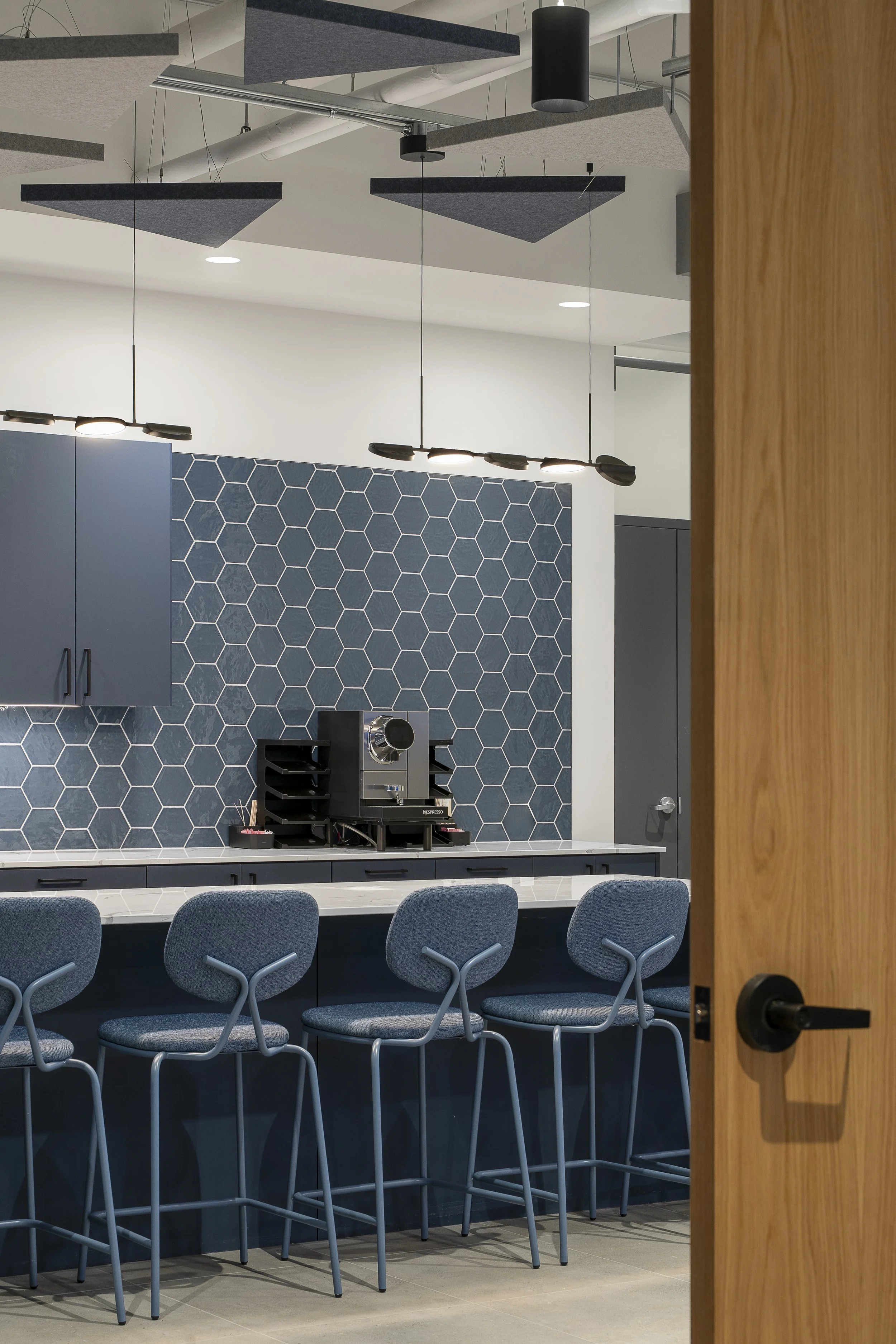 Modern cafe interior with a white countertop, four blue upholstered bar stools, blue hexagonal tile backsplash, and black pendant lighting.