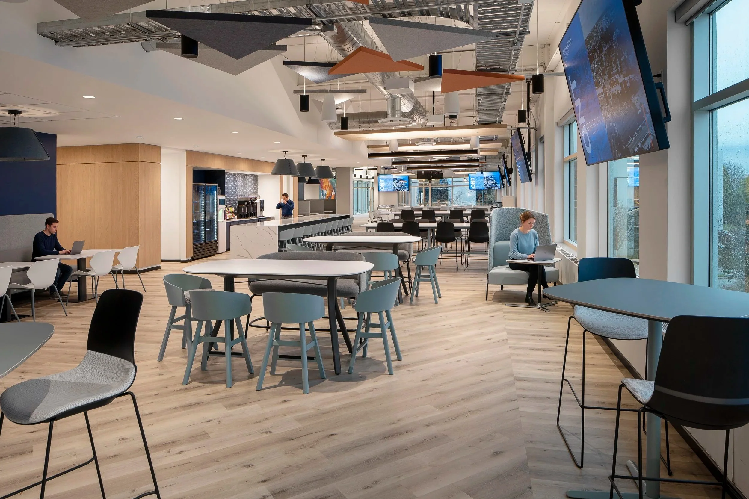 Modern cafeteria or common area with tables, chairs, and a bar area in the background, with large windows providing natural light, and multiple wall-mounted screens.