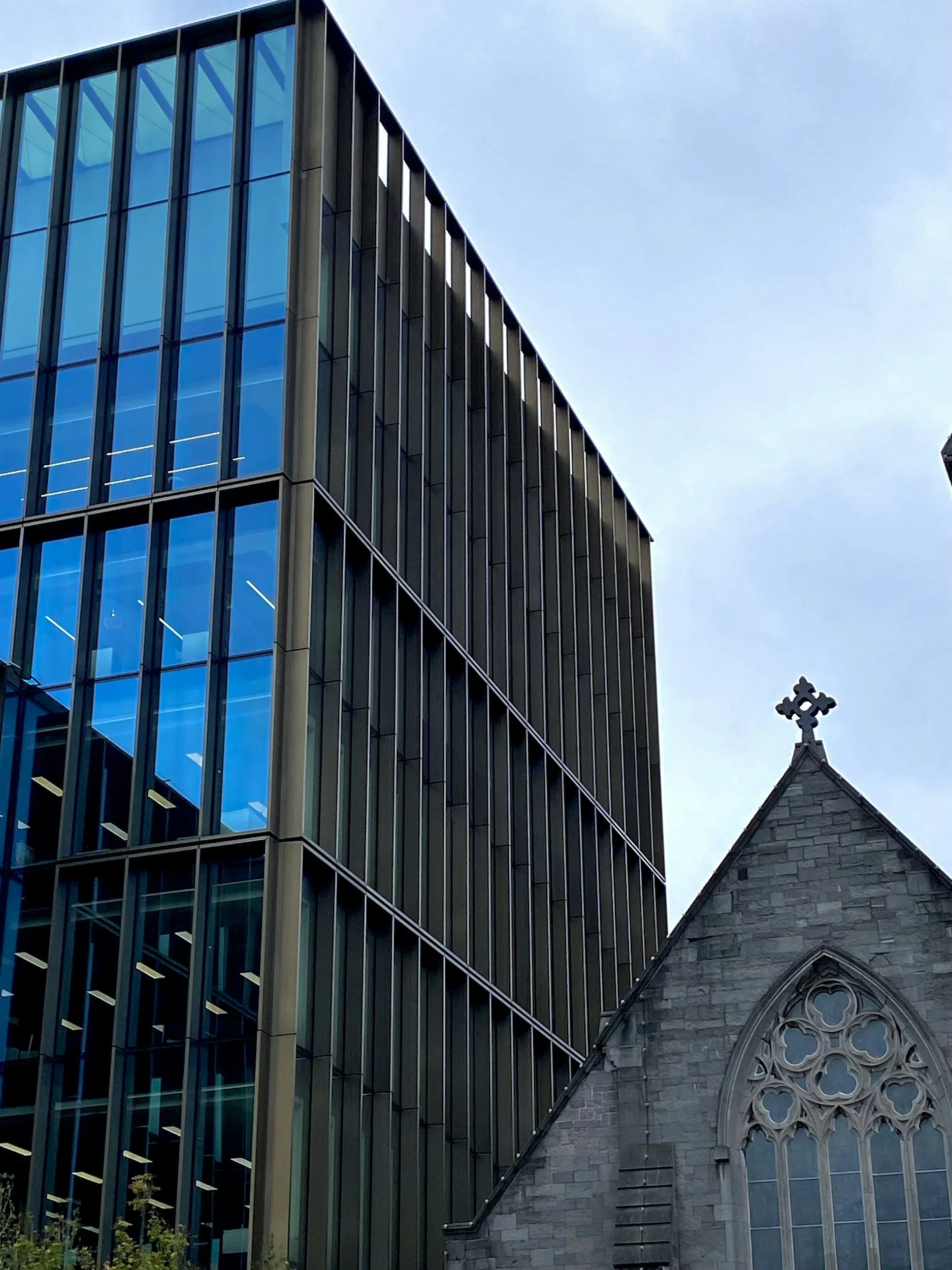 A modern building with glass windows and metal framing contrasts with an older stone church building with gothic-style arched stained glass windows and a decorative cross on the roof.