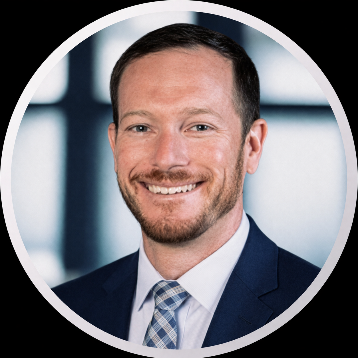 Professional headshot of a smiling man wearing a suit, tie, and dress shirt, with a blurred office background.