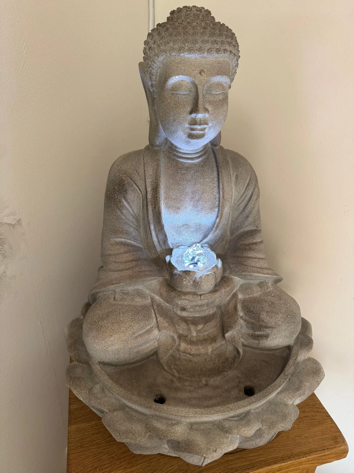 Stone Buddha statue seated with hands holding a glowing crystal ball, on a decorative circular base with a wooden surface underneath.