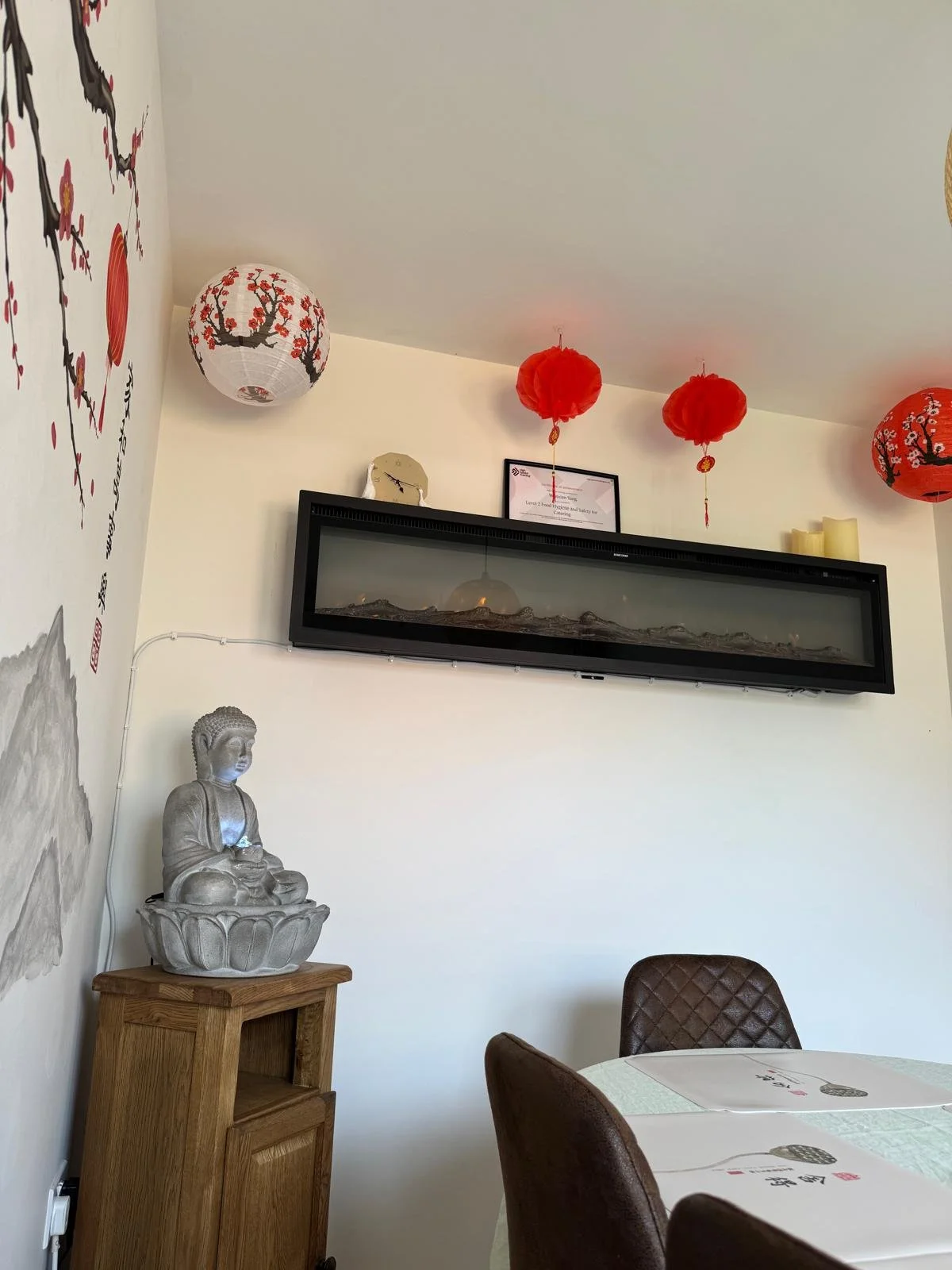 Decorative Asian-inspired interior with hanging red and white lanterns, a Buddha statue on a wooden side table, and a wall-mounted terrarium with landscape scenery.