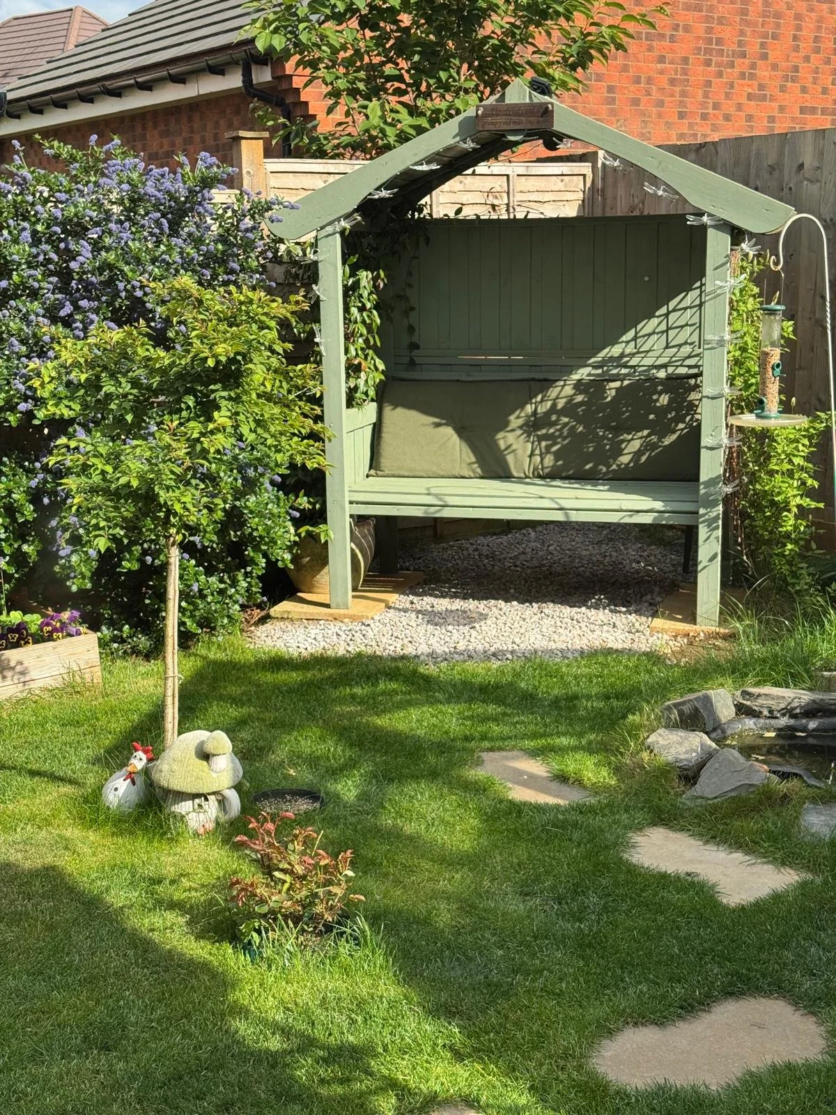 A garden scene with a green wooden outdoor swing, surrounded by flowering bushes, a small decorative mushroom ornament, small plants, and a stone pathway, with a brick house and a wooden fence in the background.
